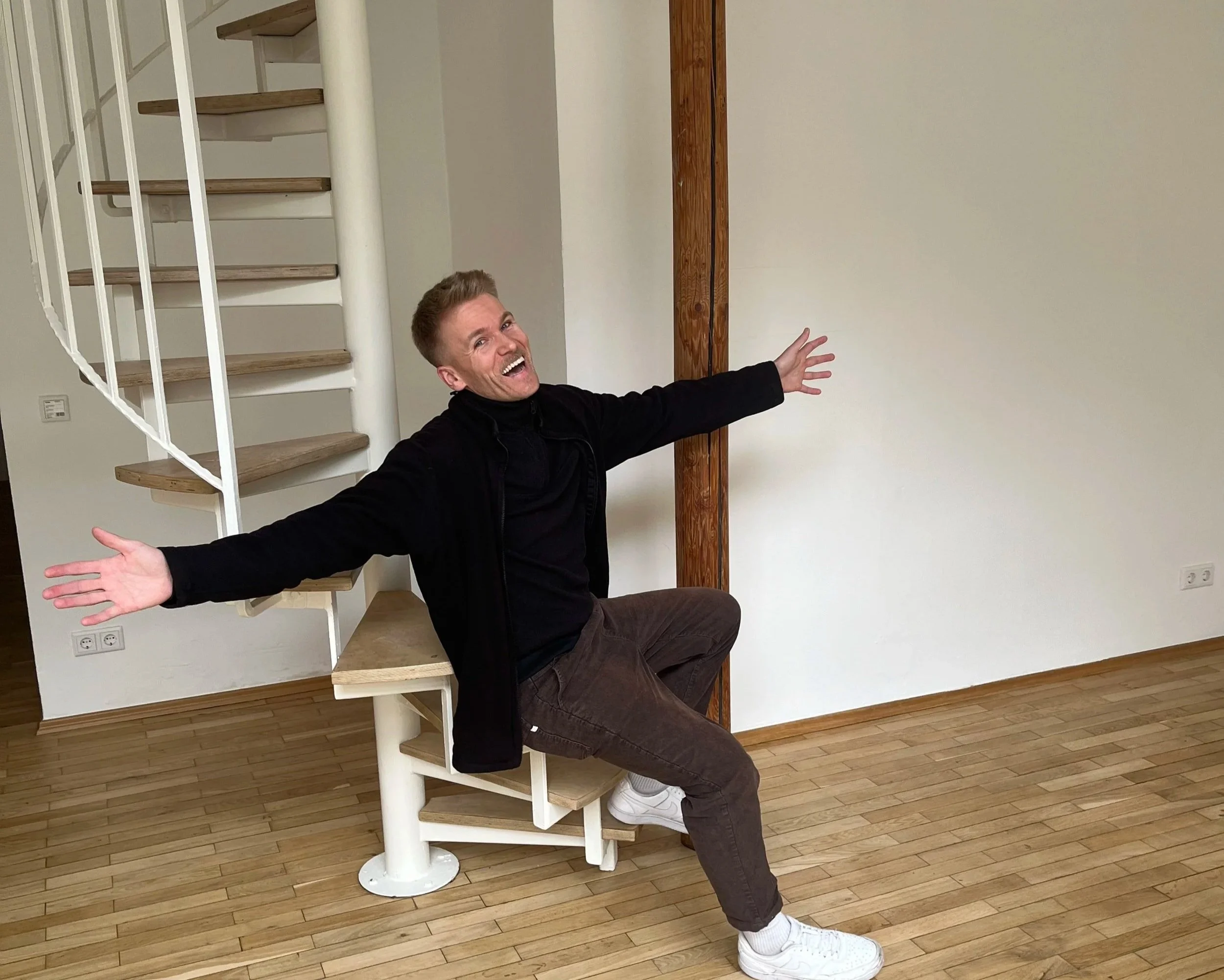 Ein lächelnder Mann sitzt auf einer Treppe in einem leeren Raum, die Arme weit ausgebreitet.