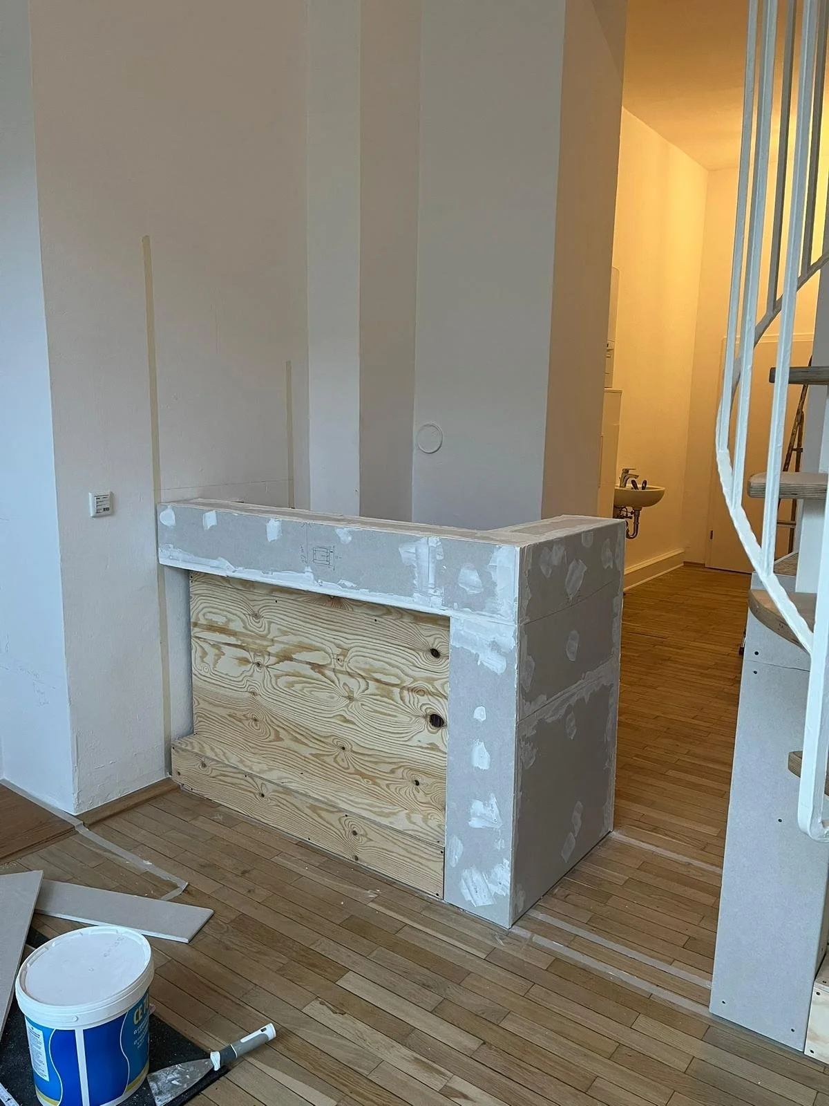 Innenaufnahme eines Hauses im Bau, mit einer halb fertiggestellten Mauer aus Gipskartonplatten und Holz. Es gibt eine kleine Spüle im Hintergrund, eine Spirale Treppe links und Bauhelferwerkzeuge auf dem Boden.