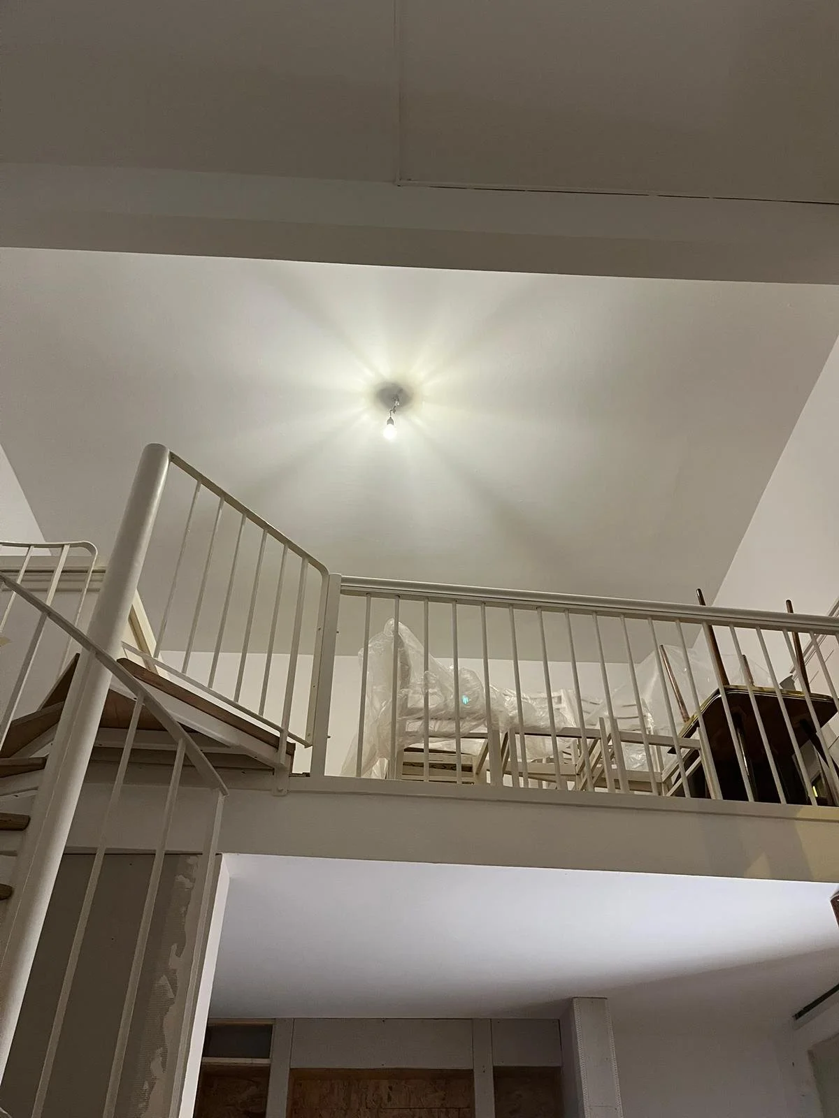 Innenaufnahme eines zweistöckigen Hauses, Blick nach oben auf eine weiße Decke mit Deckenleuchte, eine weiße Treppe auf der linken Seite, und eine offene Balkonebene mit einer Schutzbrüstung, auf der Möbel und Gegenstände stehen.