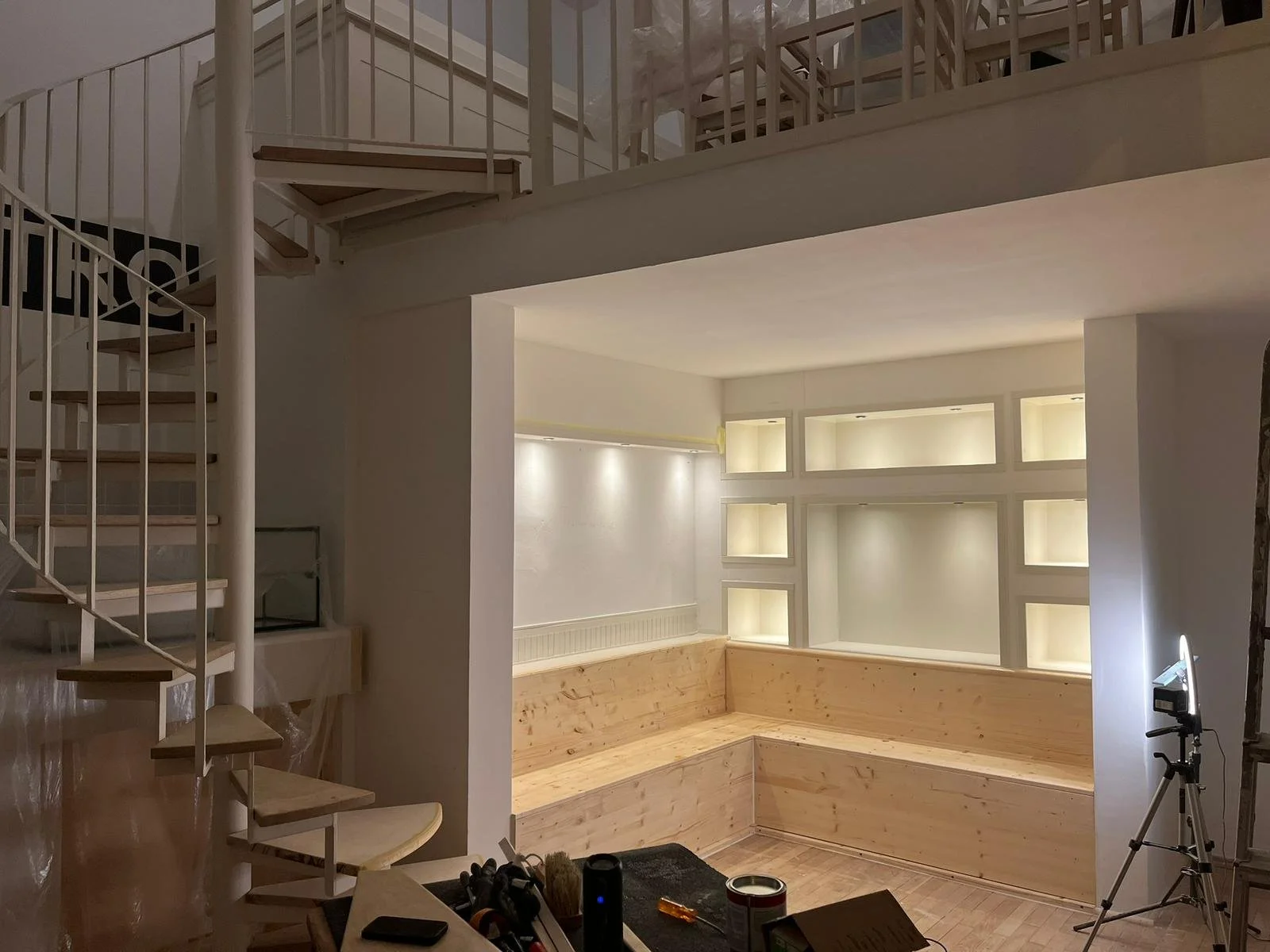 Ein heller Raum mit einer sitzbankartige Holzbank in der Ecke, beleuchtet durch eingebaute Wandlichter, und ein offenes, halb bestehendes Regal. Es gibt eine Treppe links, die nach oben führt, und eine Kamera auf einem Stativ auf der rechten Seite.