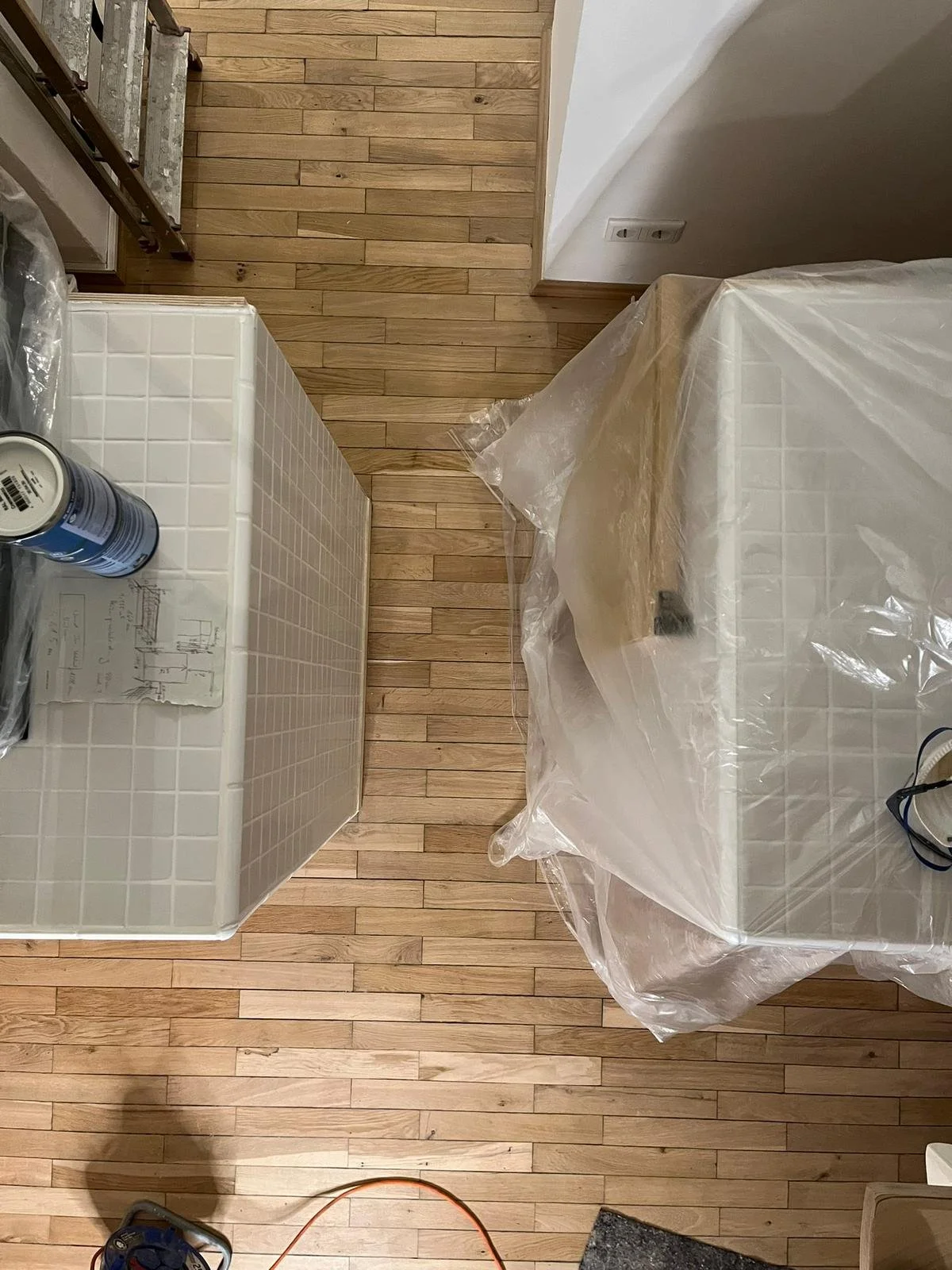 Der Raum ist im Umbau, es gibt mehrere Möbelstücke, die in Plastikfolie verpackt sind, darunter ein Tisch und eine Kommode. Es sind auch Baumaterialien und Werkzeuge sichtbar.