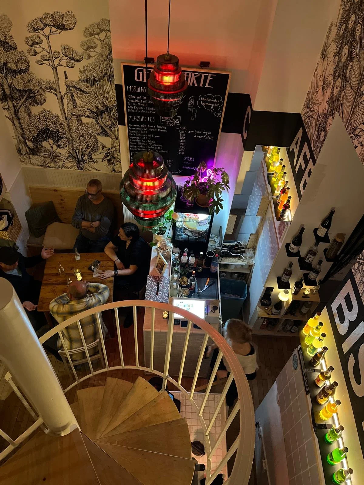 Innenaufnahme eines Restaurants oder einer Bar mit mehreren Menschen, einem Spiralaufgang, einer Theke mit Getränken, dekorativen Lampen und Wanddekorationen.