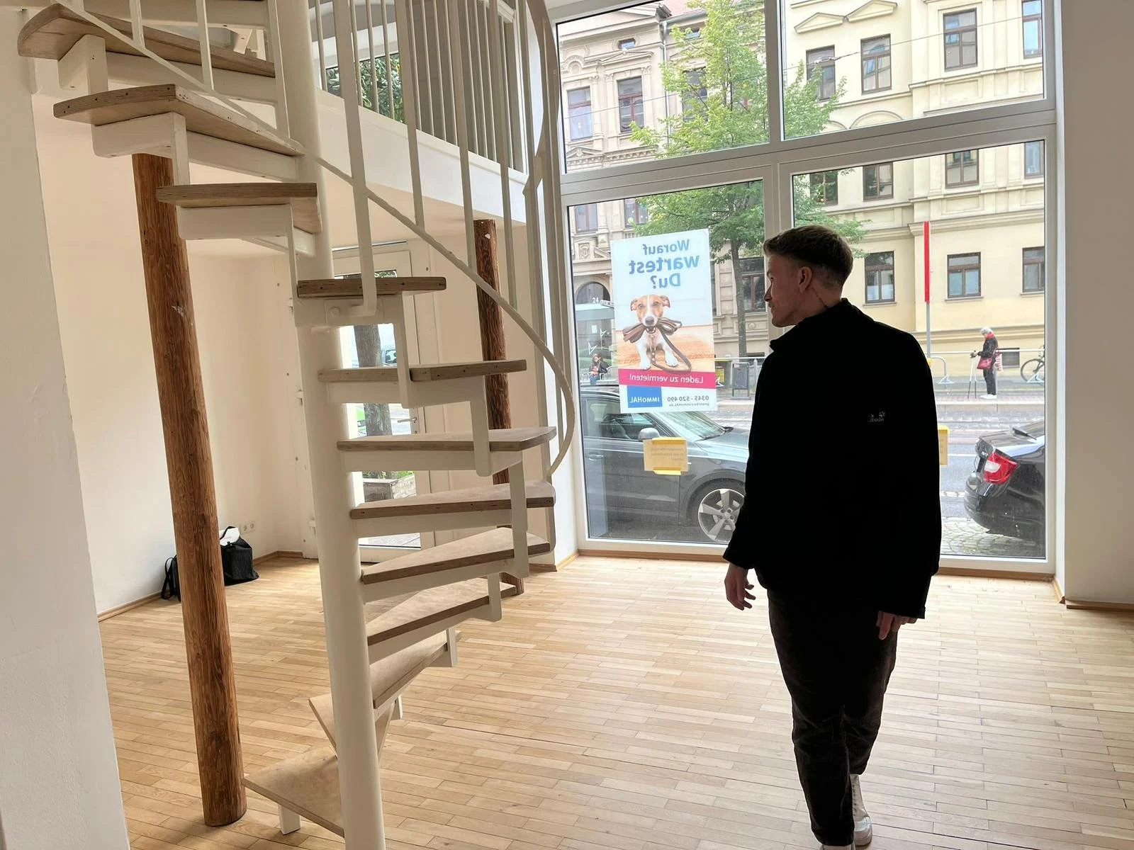 Ein Mann steht in einem leeren Raum, der durch große Fenster mit Blick auf die Straße sichtbar ist. Der Raum hat einen Holzfußboden und eine weiße Wendeltreppe.