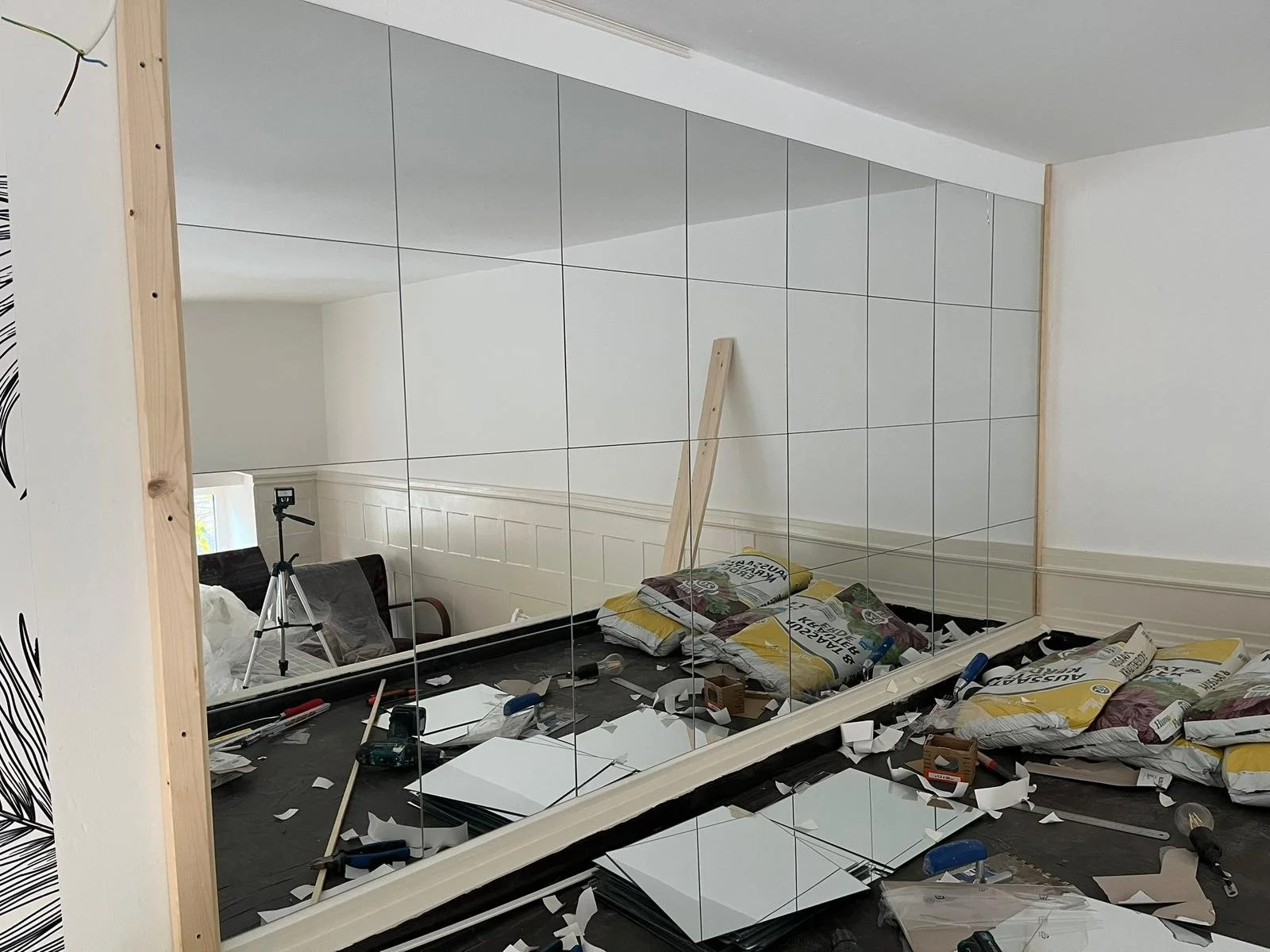 Ein Raum im Bau oder bei der Renovierung, mit einer großen Spiegelwand, zahlreichen Baumaterialien, Werkzeugen, Packungen und zerschnittenen Spiegelstücken auf dem Boden, sowie einer Holzlatte, die an der Wand lehnt.