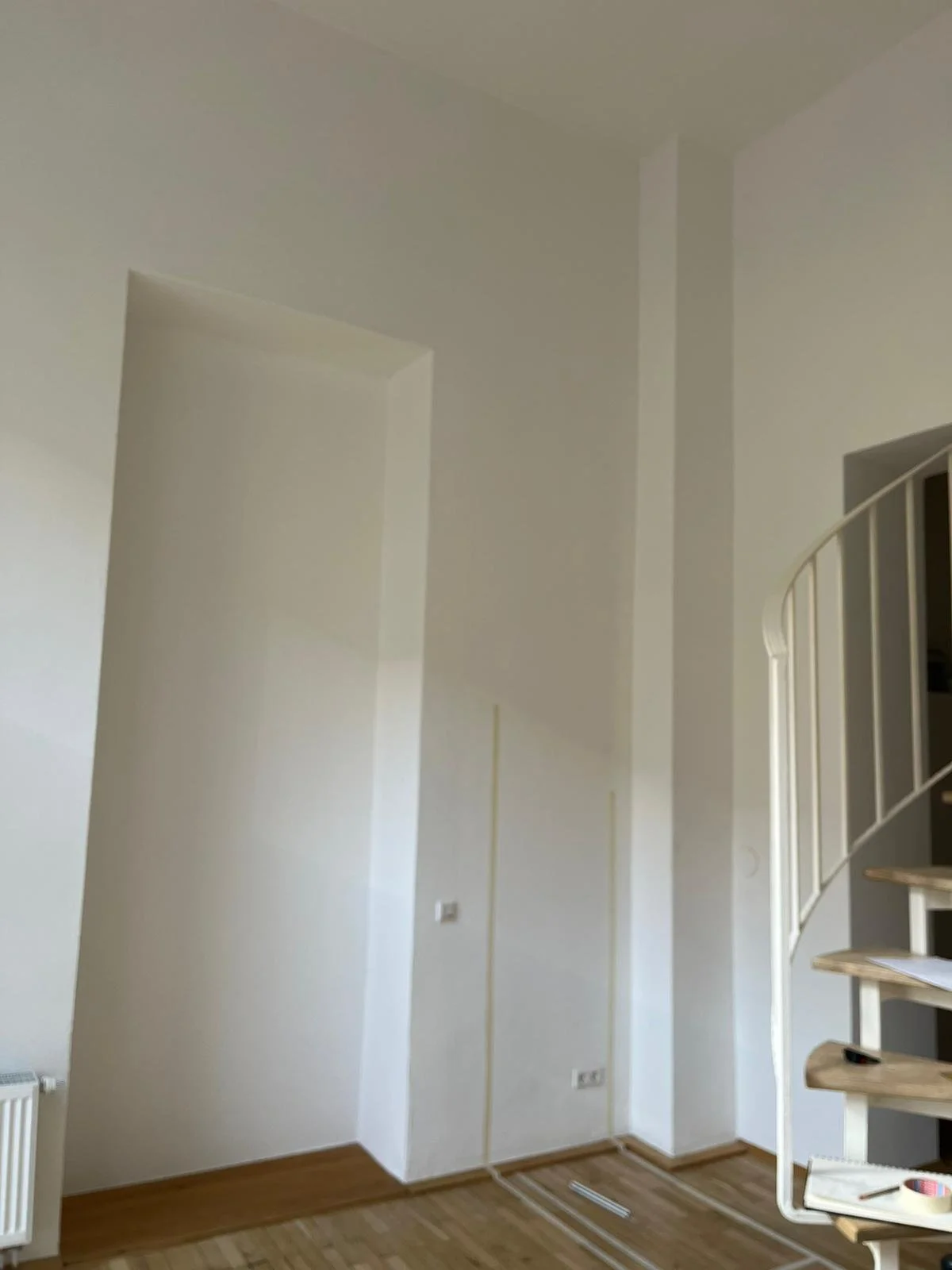 Innenaufnahme eines leeren, weißen Zimmers mit Holzboden, einer Heizkörper an der Wand, Kabel und Steckdosen auf dem Boden, und einer Treppe mit weißen Geländer auf der rechten Seite.