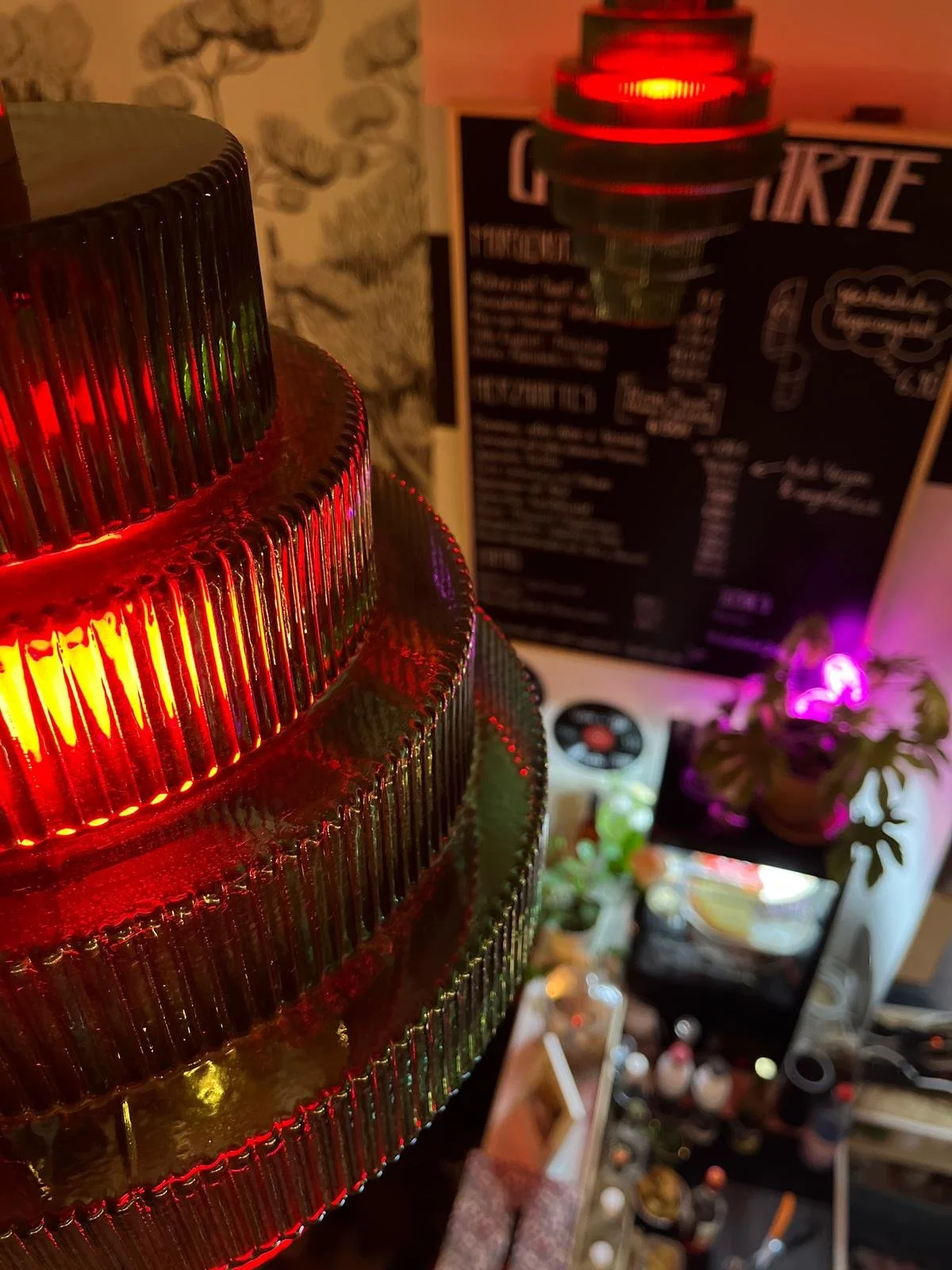 Blick auf eine mehrstöckige, gläserne Lampe mit roten Lichtreflexen im Vordergrund. Im Hintergrund sind eine Tafel mit Speisekarte, Pflanzen mit bunten Lichtern und verschiedene Dekorationen sichtbar.