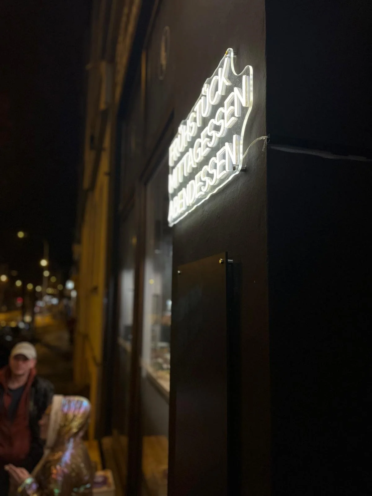 Leuchtendes Neon-Schild an einer schwarzen Wand, nachts, draußen. Im Hintergrund sind Straßenlaternen und unscharfe Personen und Fahrzeuge zu sehen.