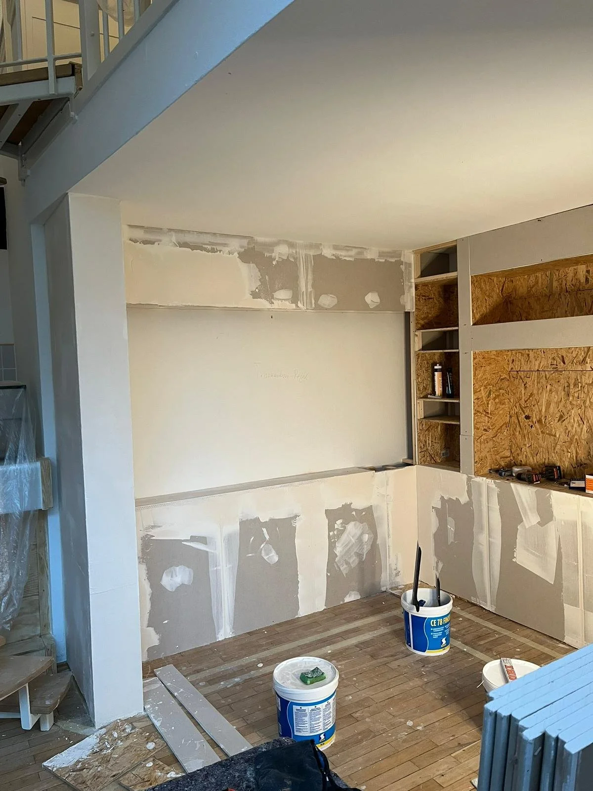 Innenraum eines Hauses beim Renovieren. Es sind Putzreste an der Wand, Bauwerkzeuge und Material im Raum verteilt, inklusive Eimer und Holzplatten.
