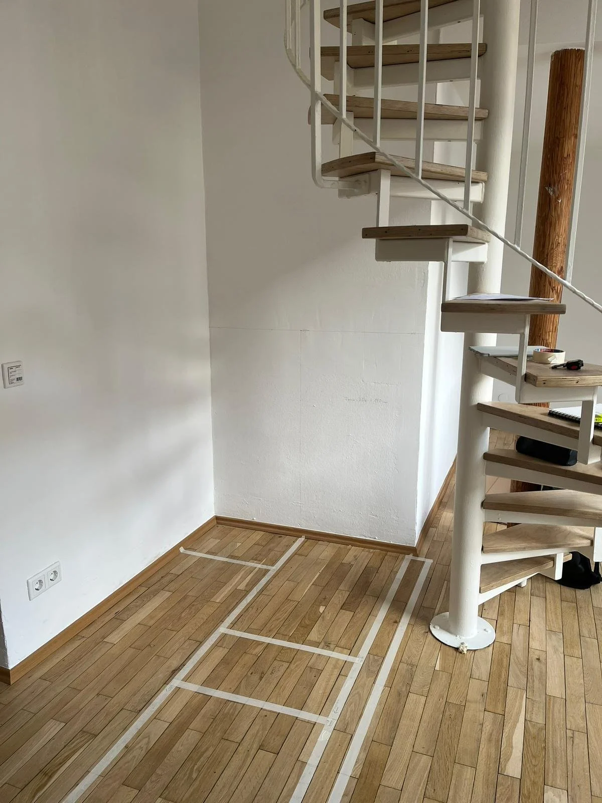 Leerer Raum mit Holzfußboden, markierten Bereichen auf dem Boden, einer weißen Wendeltreppe und einer Holzstützsäule.