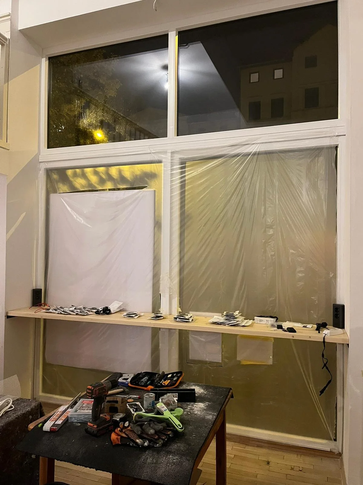 Innenraum eines Zimmers oder Balkons in der Renovierung, mit Schutzfolie an den Fenstern, einem Arbeitstisch mit Werkzeugen und Materialien, sowie div. Elektrokabeln und Ausrüstungsgegenständen.