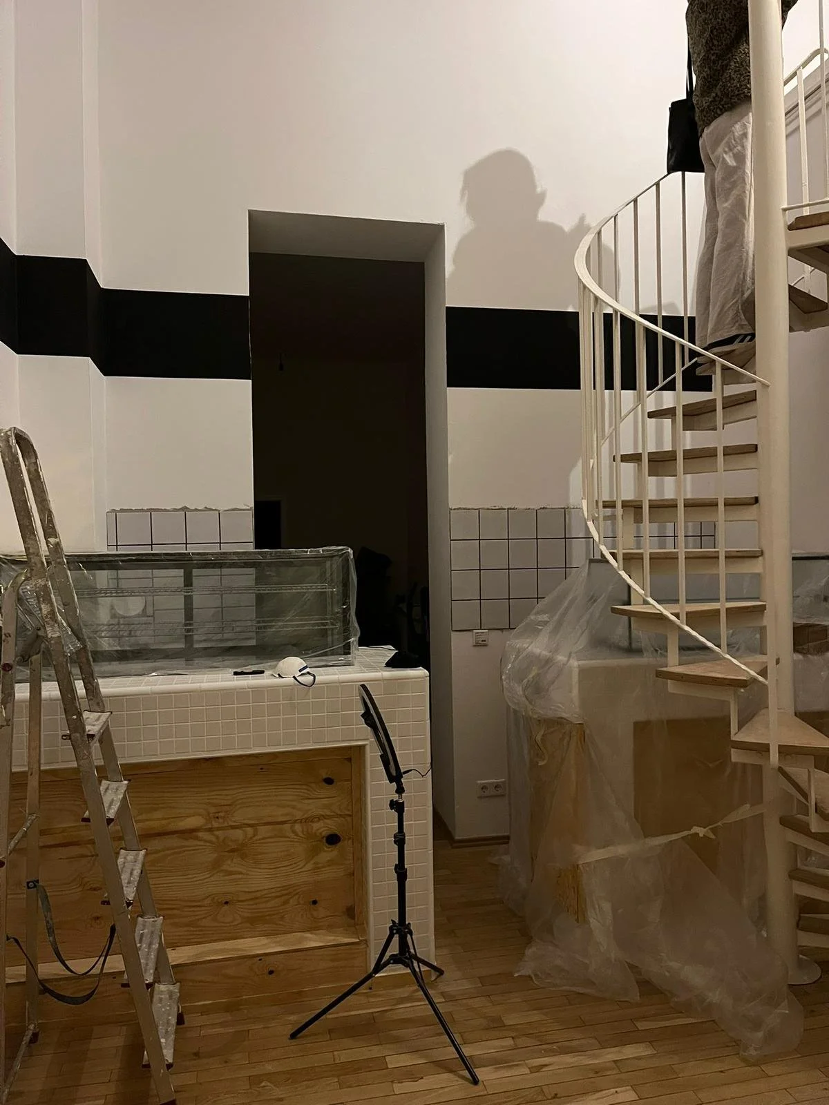 Innenaufnahme einer Baustelle mit Leiter, Stativ, Holz, Fliesen, Treppe und einer Person, die an der Treppe arbeitet.