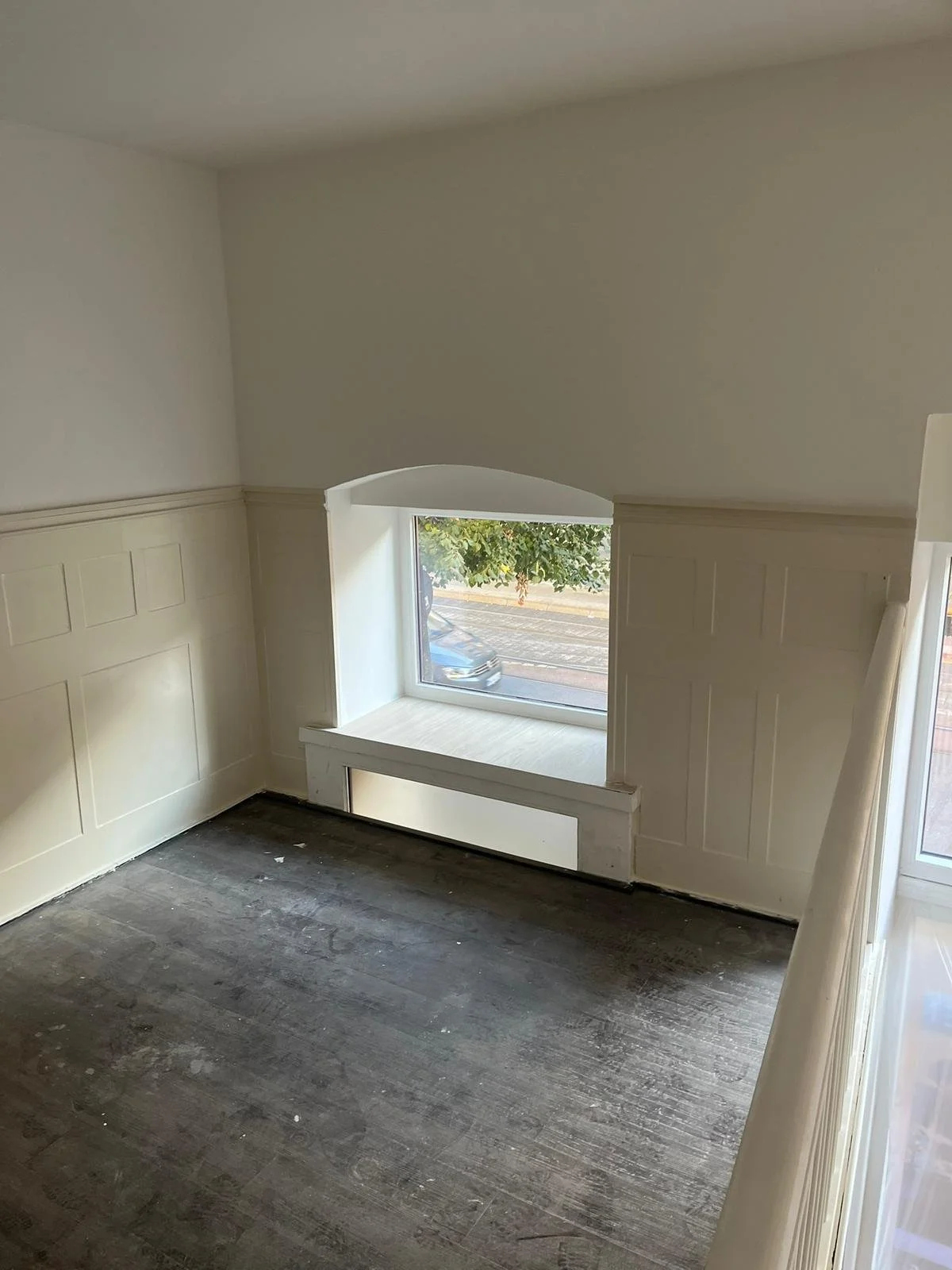 Ein leerer Raum mit weißen Wänden, dunklem Holzboden und einem großen Fenster, durch das eine Straße mit einem abgestellten Auto zu sehen ist.