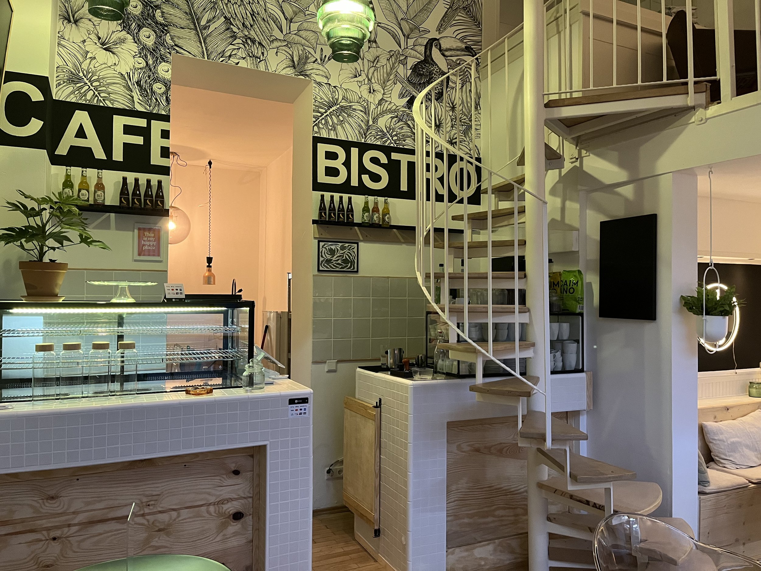 Innenansicht eines Cafés mit einer Treppe, die in eine zweite Etage führt, eine Wand mit großen schwarzen Buchstaben, die 'CAFE' und 'BISTRO' sagen, und dekorativer Wandmalerei mit tropischen Pflanzen und einem Vogel.