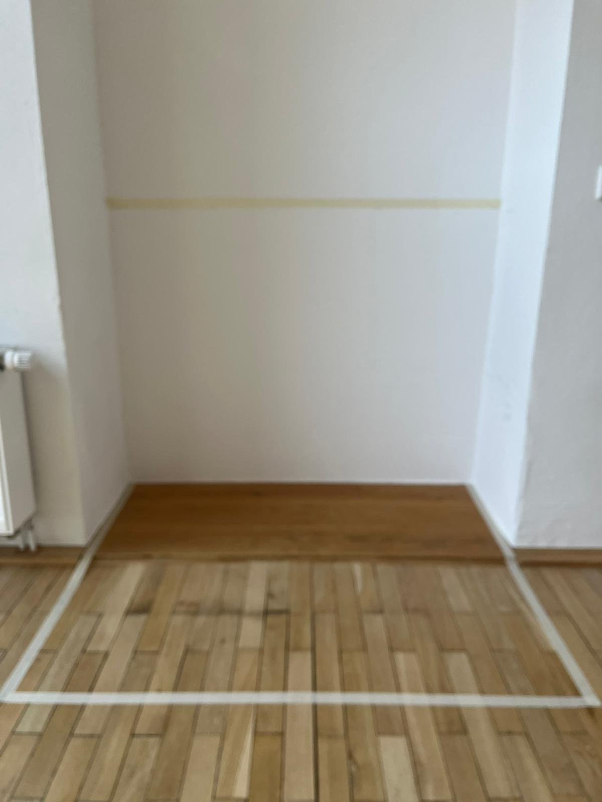 Leerer Raum mit weißen Wänden, braunem Holzboden und einer Markierung auf dem Boden.