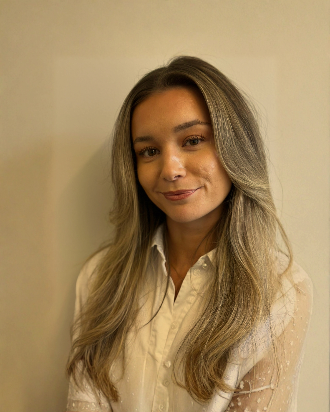 A woman with long, wavy light brown hair wearing a white blouse, standing against a neutral beige wall, smiling softly.