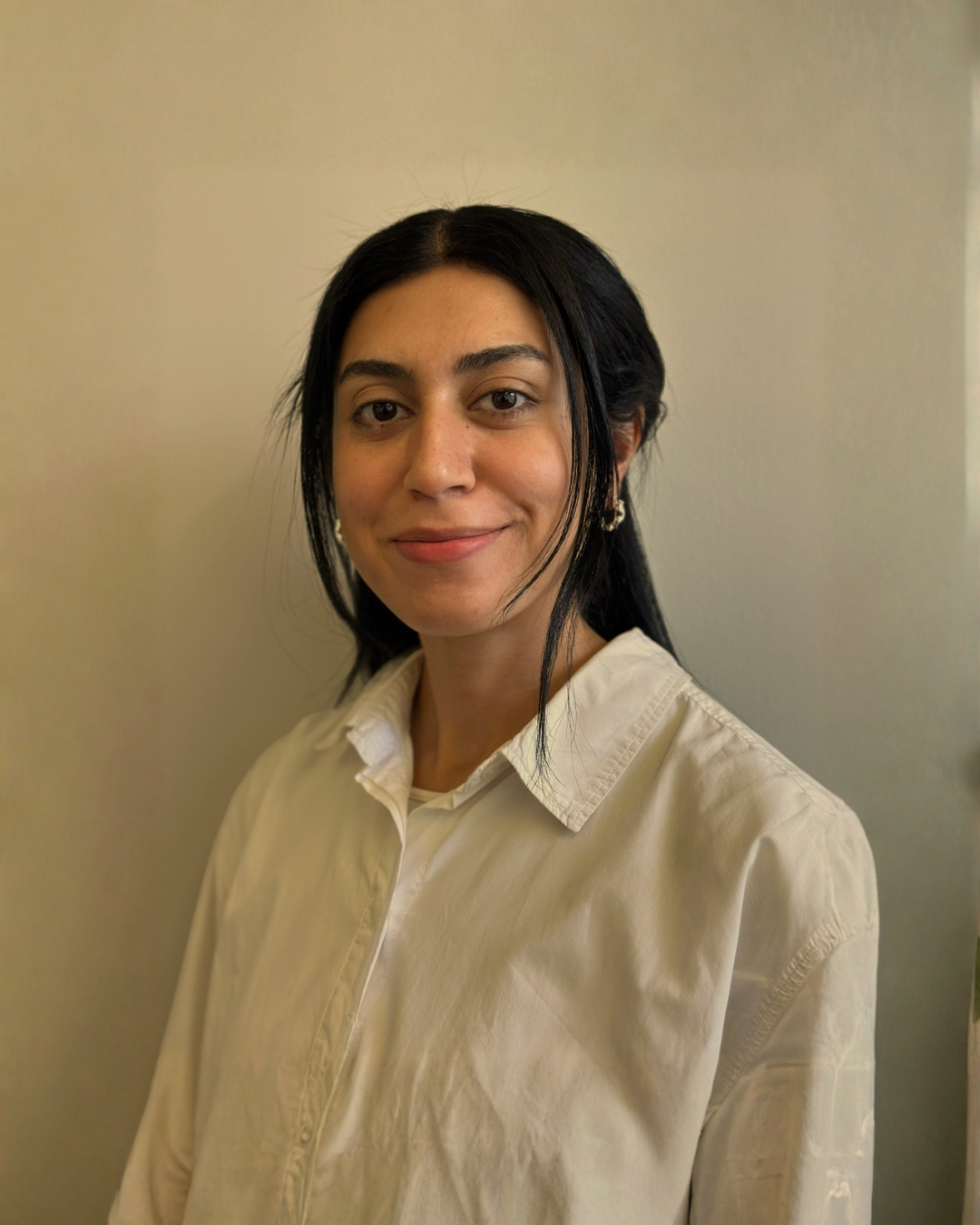 A woman with black hair, wearing a cream-colored button-up shirt, standing against a neutral wall.