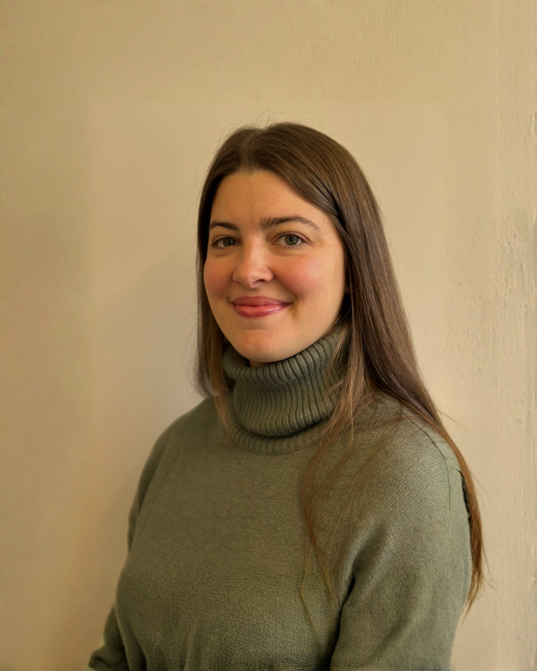 A woman with long brown hair wearing a green turtleneck sweater, standing in front of a plain, off-white wall.