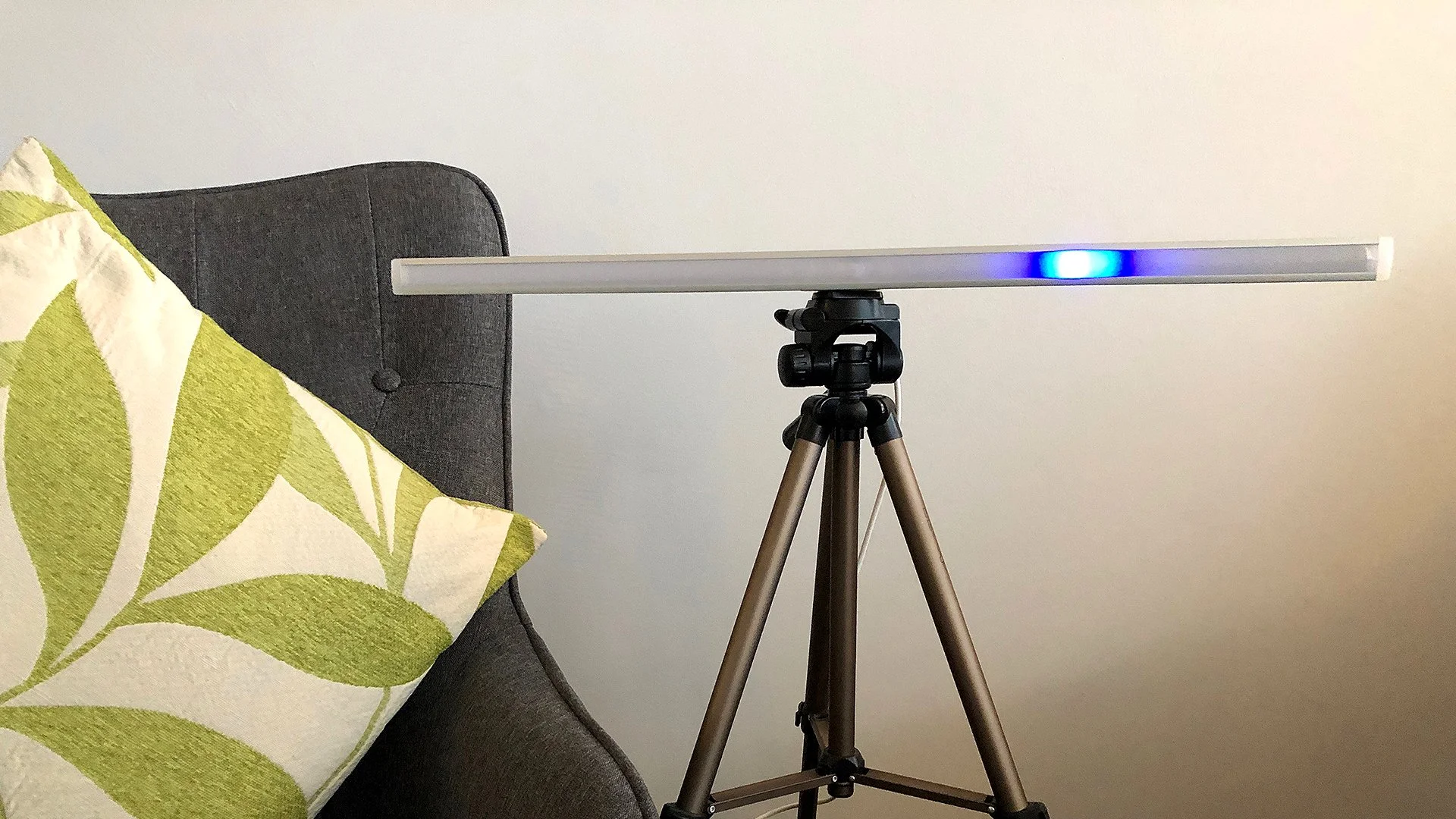 A tripod with a long, rectangular light bar mounted on top, positioned in front of a gray sofa with a green and white patterned pillow, against a plain white wall.