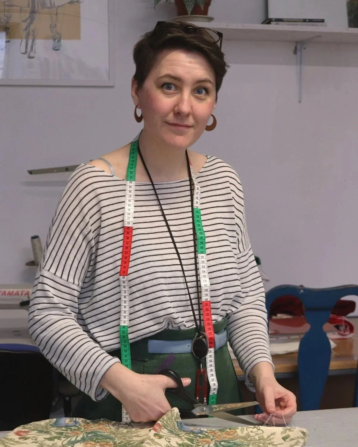 A woman with short brown hair, wearing a striped shirt and glasses on her head, is standing at a work table, holding scissors to cut fabric. She has a measuring tape draped around her neck and is smiling at the camera.