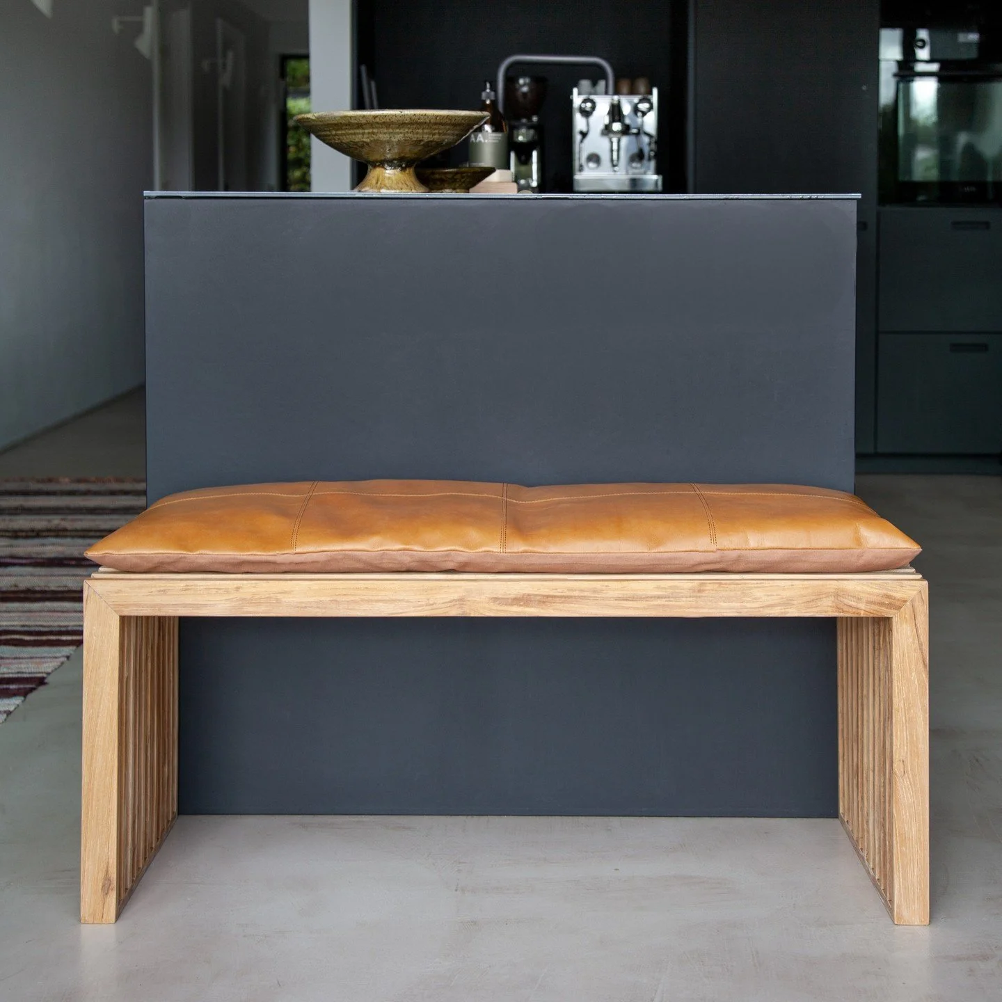 A modern kitchen with a wooden bench with a tan leather cushion in front of a dark gray kitchen island, with a decorative bowl on top and kitchen appliances in the background.