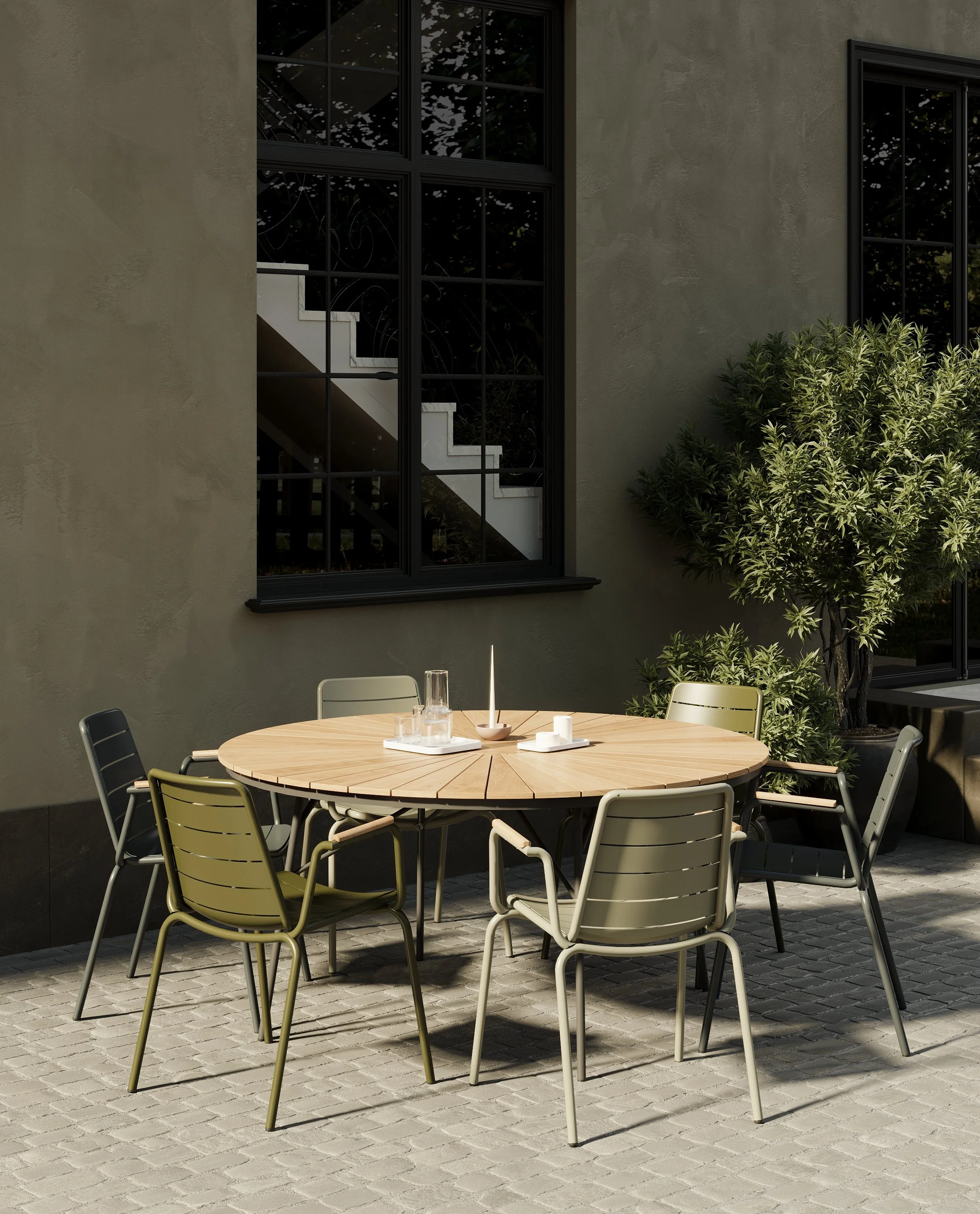 Outdoor patio with a round wooden table and six chairs, set against a gray wall with large windows, and a potted bush to the right.