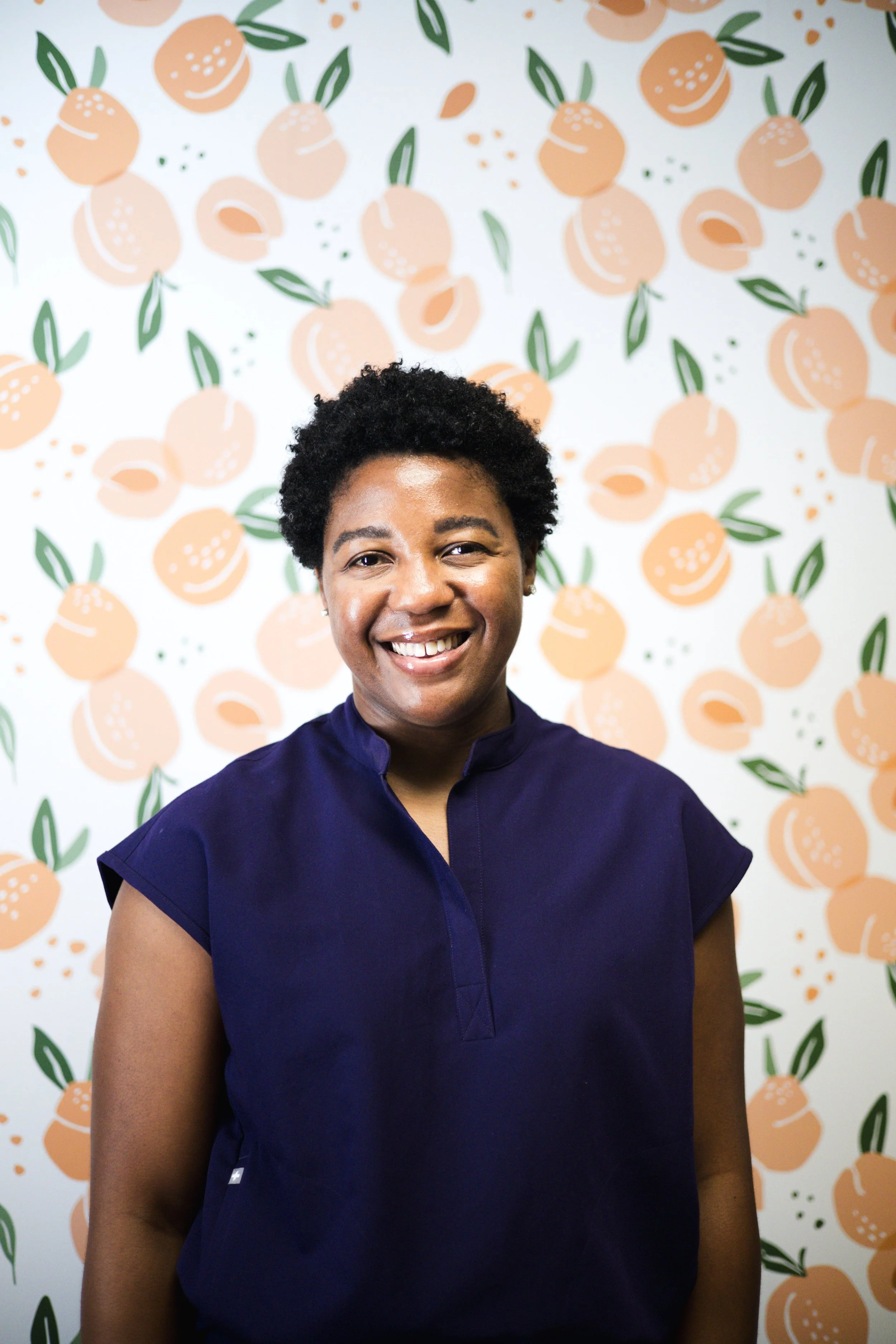Portrait of a smiling woman with short curly black hair, wearing a navy blue top, standing in front of a patterned orange and green fruit-themed background.
