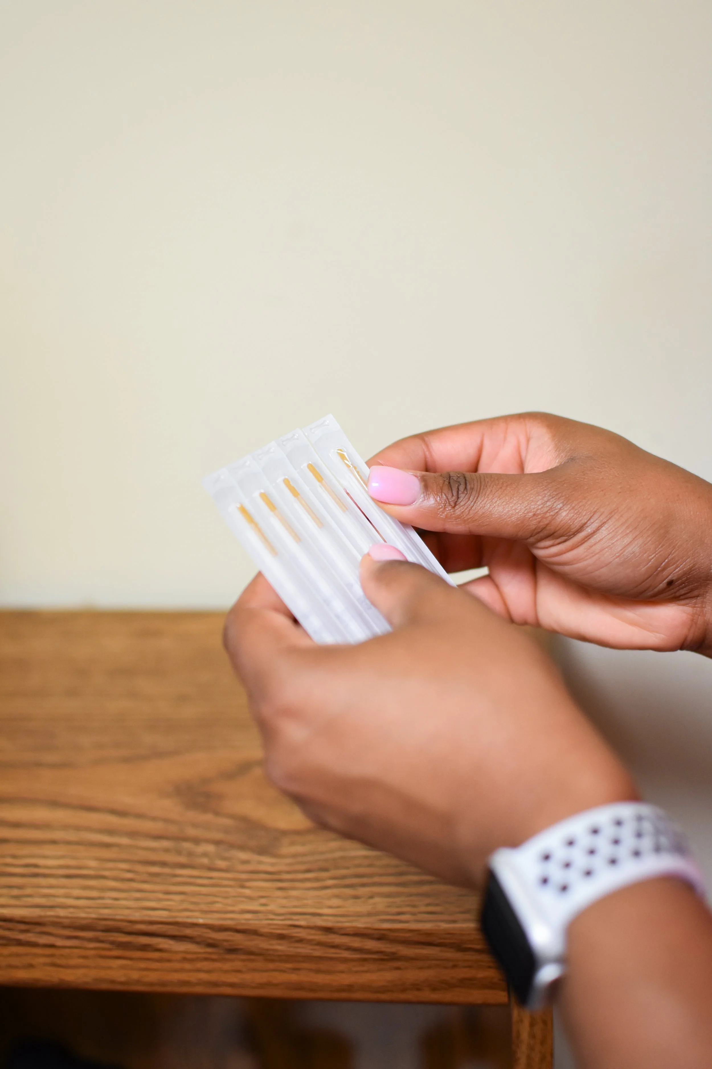 A person with dark skin and pink nail polish holding a box of pregnancy test kits with a beige wall background.