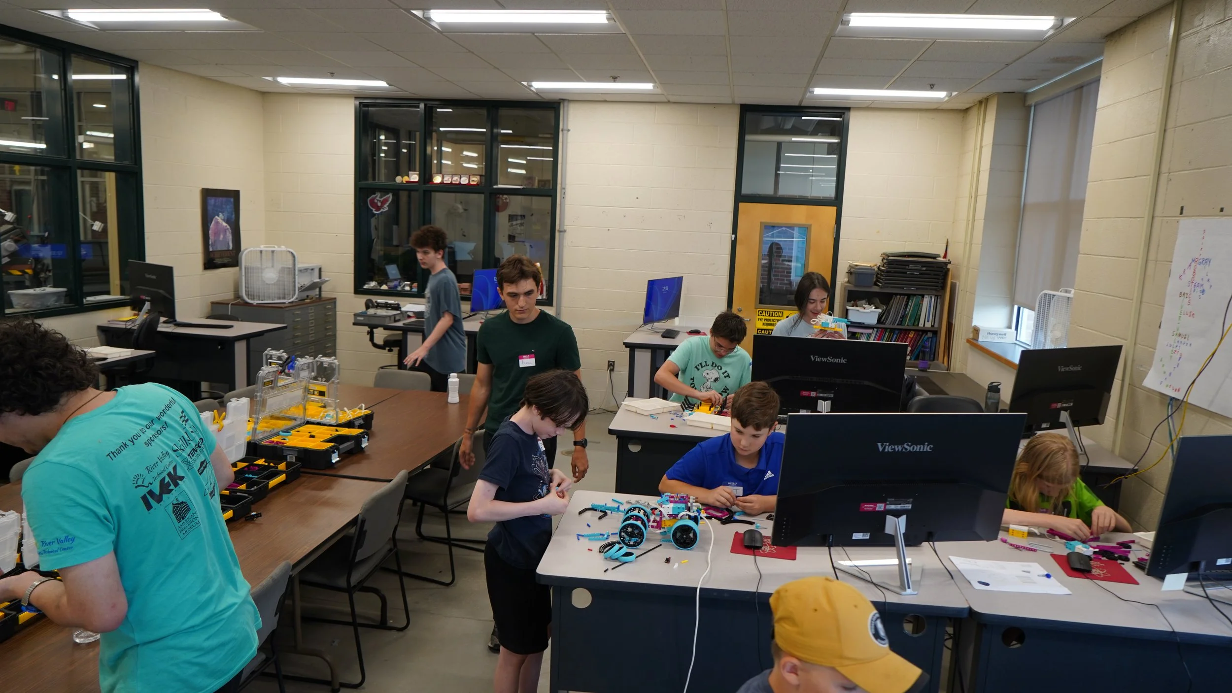 Children and teenagers working on robotics projects at a classroom table, assembling and programming robots with tools, computers, and electronic components.