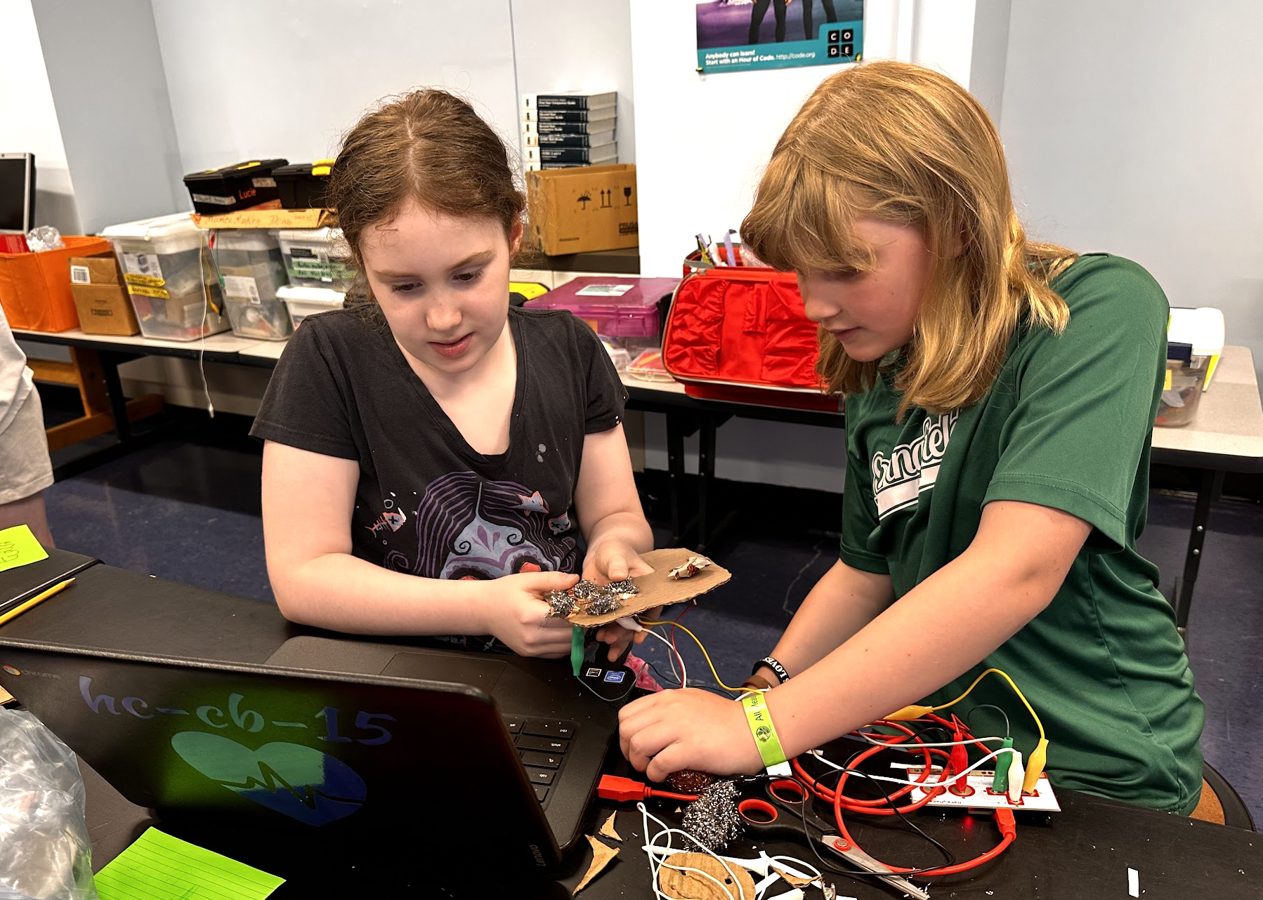 Two girls working on an electronics project at a workshop or classroom, with wires, tools, and a laptop on the table.