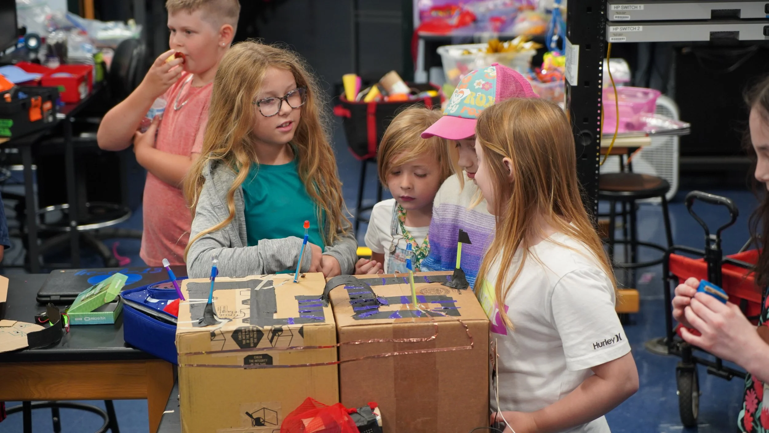 A group of children gathered around a cardboard project with various decorations, observing and discussing it in a classroom or workshop setting.