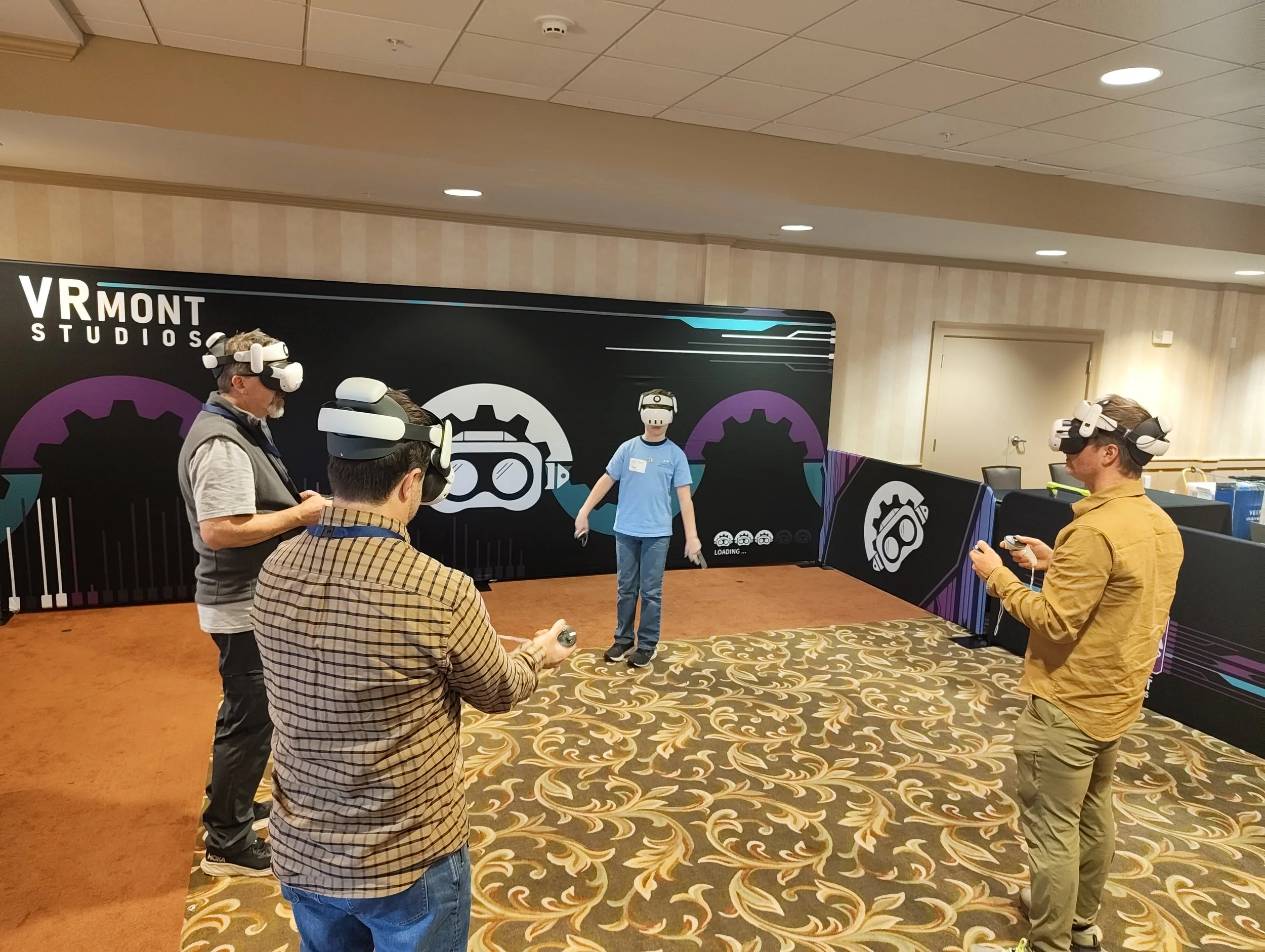 Group of four people using VR headsets at VR Mont Studios event, one person standing on stage with VR headset, others holding controllers, decorated with VR-themed graphics.