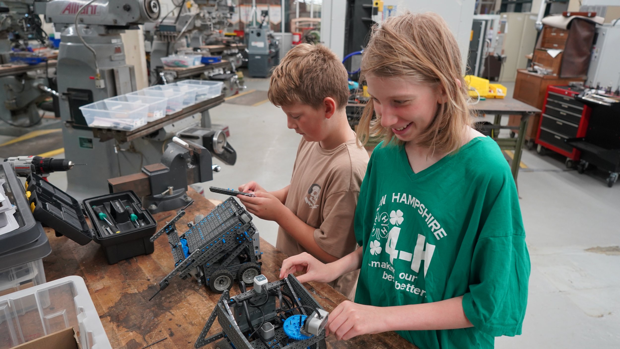 Two children working on assembling robots in a workshop or classroom with tools and equipment on a workbench.