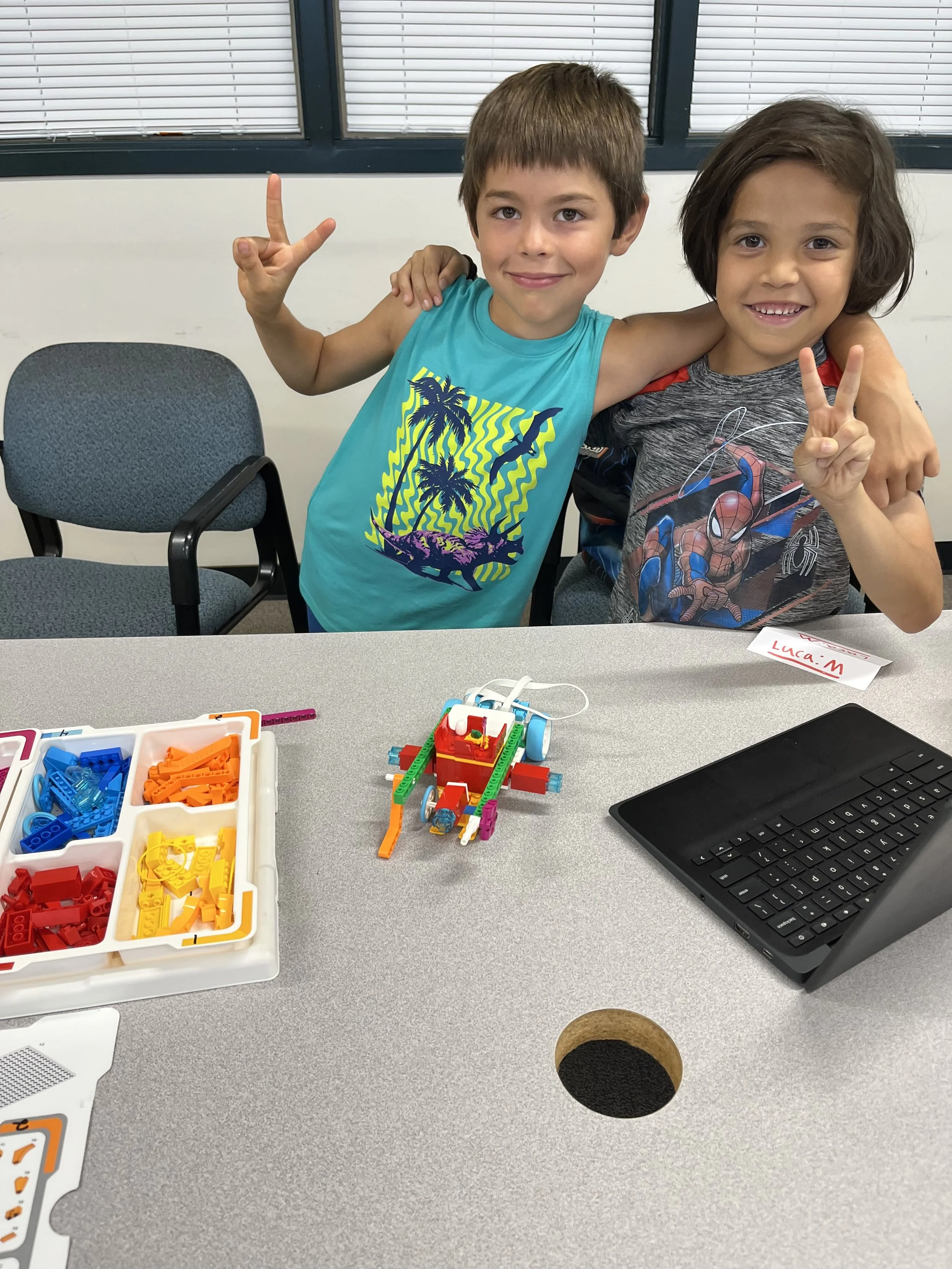 Two young boys wearing casual T-shirts show victory and peace signs, standing behind a table with a small LEGO robot and a container of colorful LEGO pieces, in a room with window blinds.