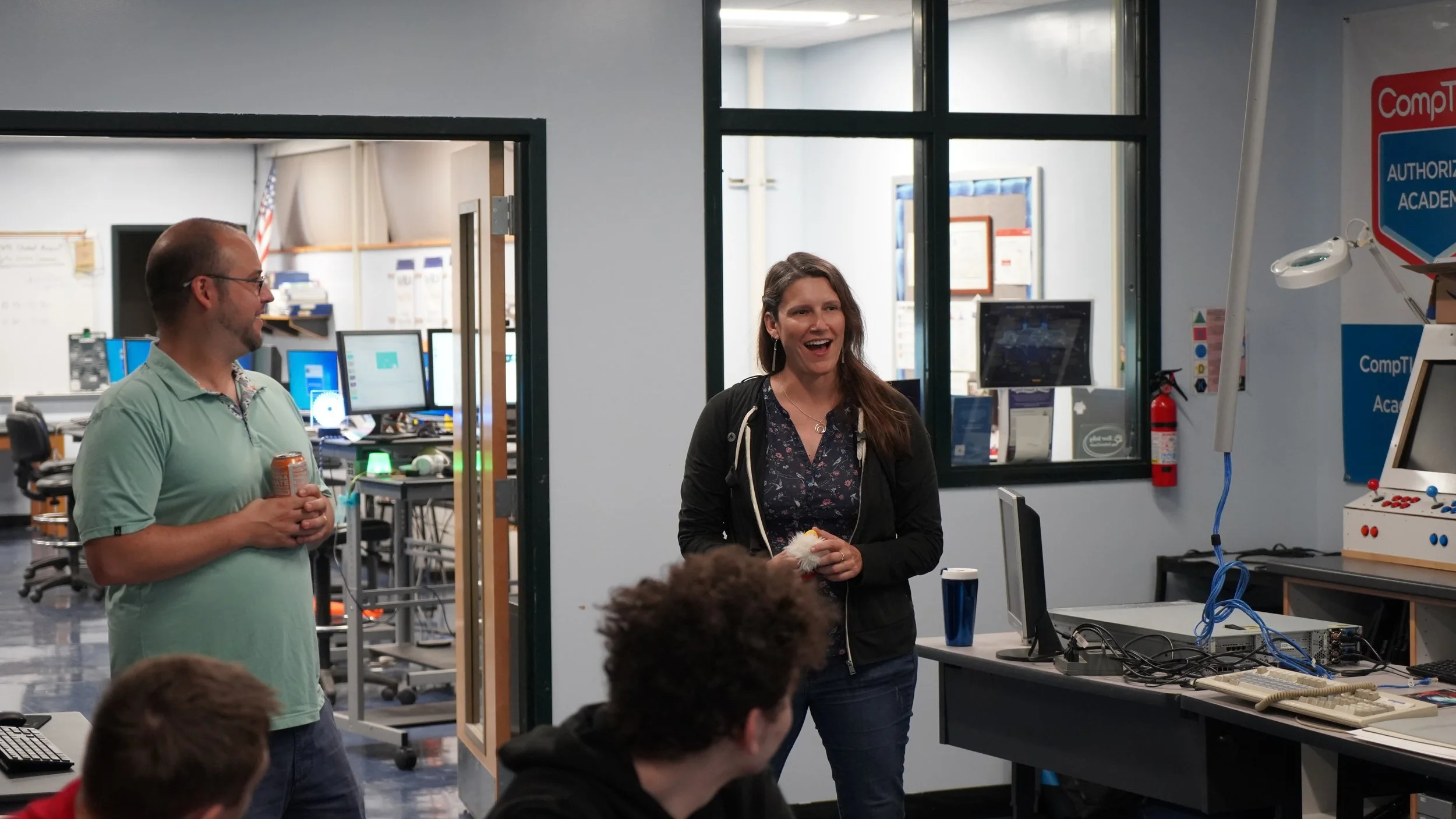 A woman talking and gesturing inside a computer lab or classroom, with a man holding a can of soda to her left, and several students seated nearby. Multiple computer monitors and classroom equipment are visible in the background.
