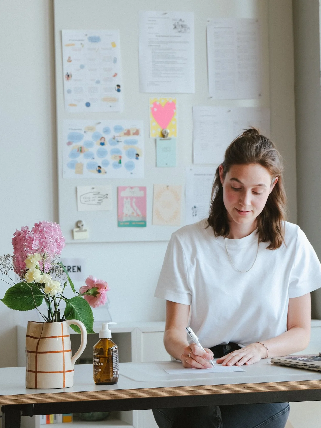 Eine junge Frau sitzt an einem Tisch, schreibt auf einem Blatt Papier, neben ihr eine Vase mit pinken und weißen Hortensien, und ein Desinfektionsmittel auf dem Tisch. Im Hintergrund ist eine Pinnwand mit verschiedenen Dokumenten und Postern.