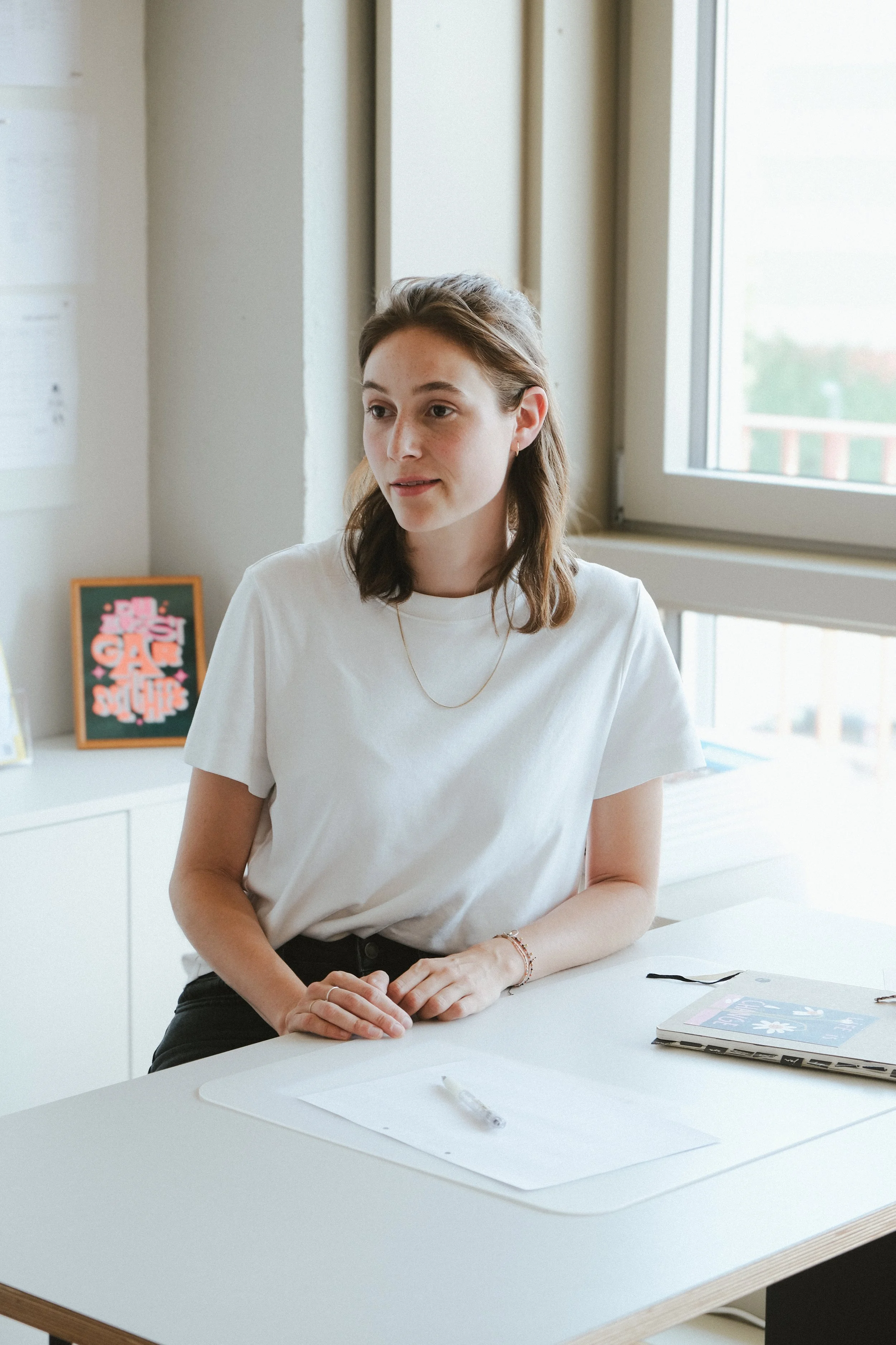Junge Frau sitzt an einem weißen Tisch in einem hellen Raum, trägt ein weißes T-Shirt, hat braune Haare, gesteigertes Interesse, es liegt ein Notizbuch und ein Stift auf dem Tisch.