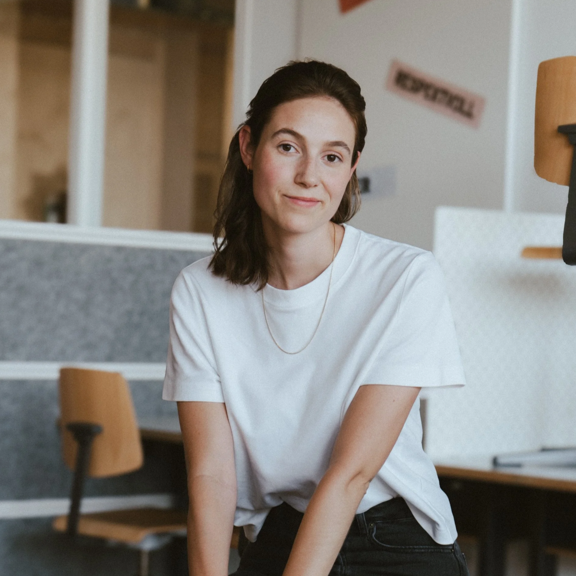Porträt einer jungen Frau mit braunen Haaren, weißen T-Shirt, in einem modernen Raum, Sitzen an einem Tisch.