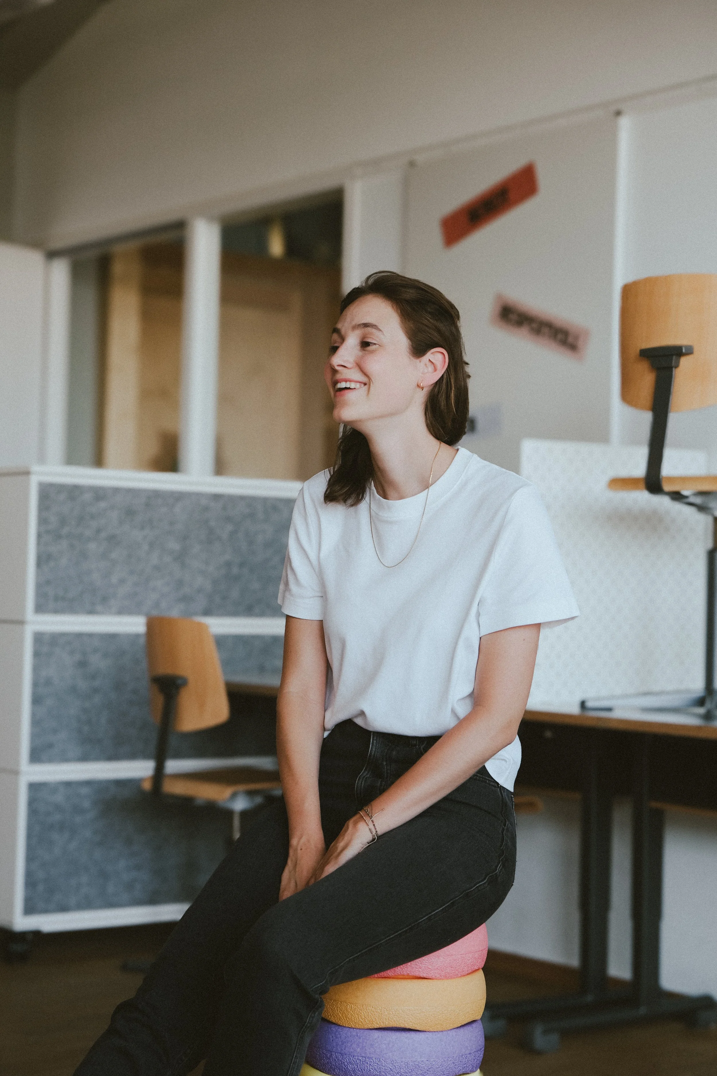 Eine Frau sitzt auf einem bunten Hocker in einem modernen Büro. Am Kopf des Hockers ist eine farbige Matte. Im Hintergrund sind weiße Wände mit roten Schildern und Büromöbeln.