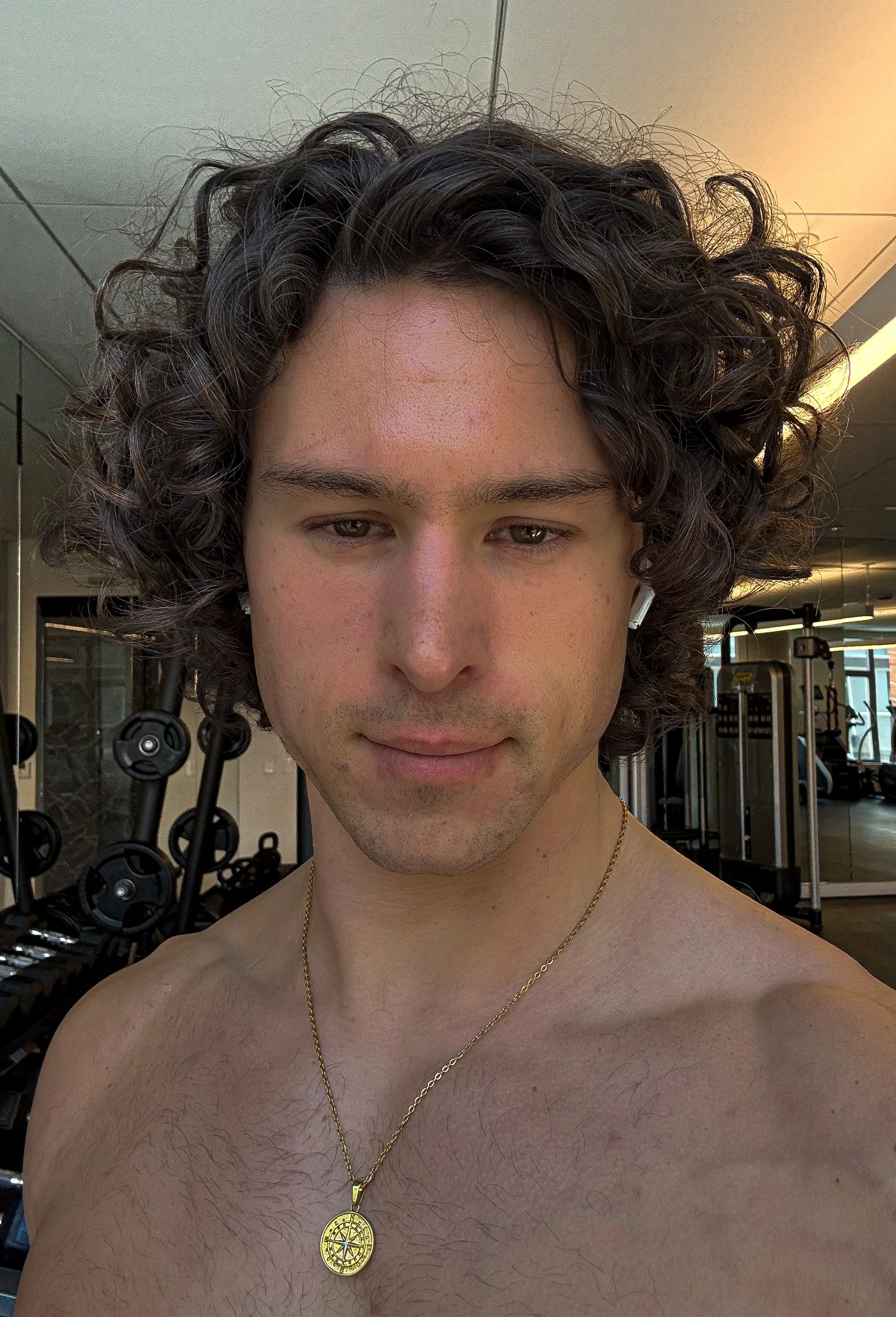 A man with curly dark hair and light skin taking a selfie shirtless in a gym, wearing a gold pendant necklace, with gym equipment visible in the background.