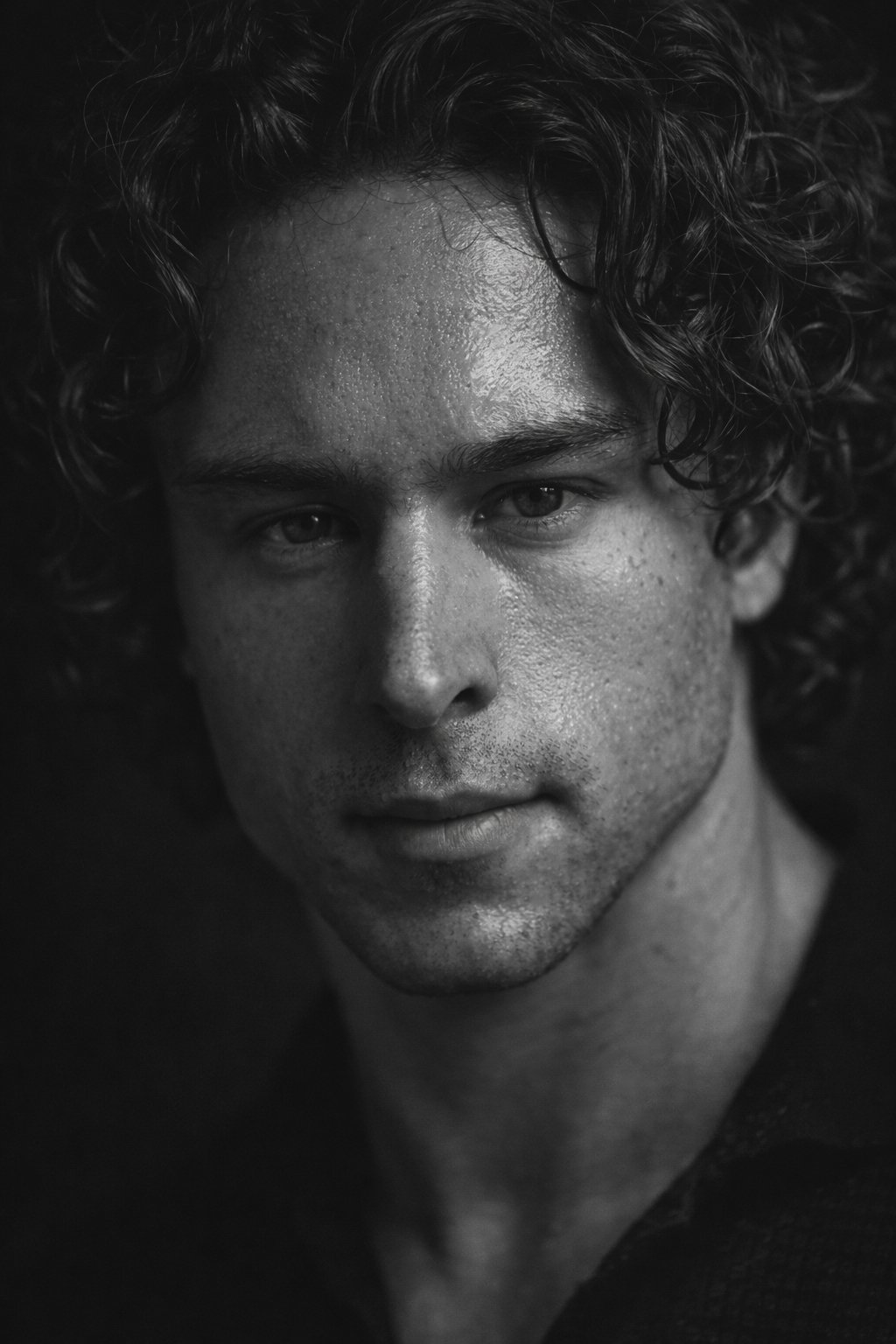 Close-up black-and-white portrait of a young man with curly hair and textured skin, looking directly at the camera.