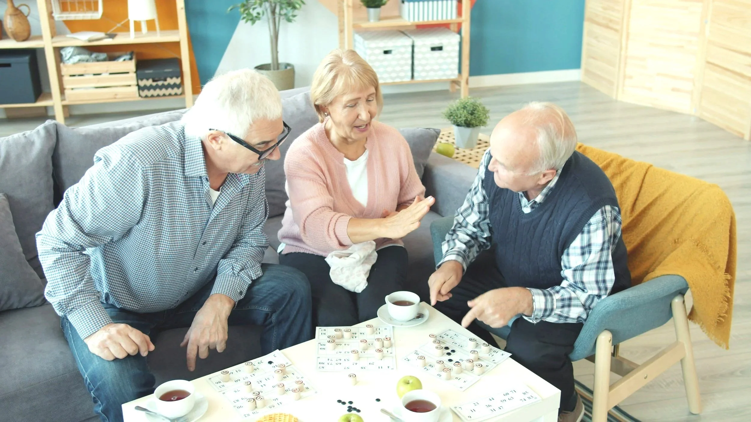 Three elderly people playing bingo and chatting in a cozy living room with a coffee table, drinks, and snacks.