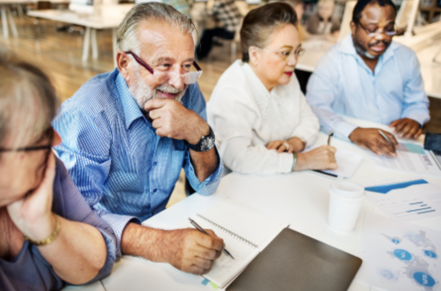 A diverse group of four people sitting at a table in a business meeting, taking notes and discussing charts and graphs.