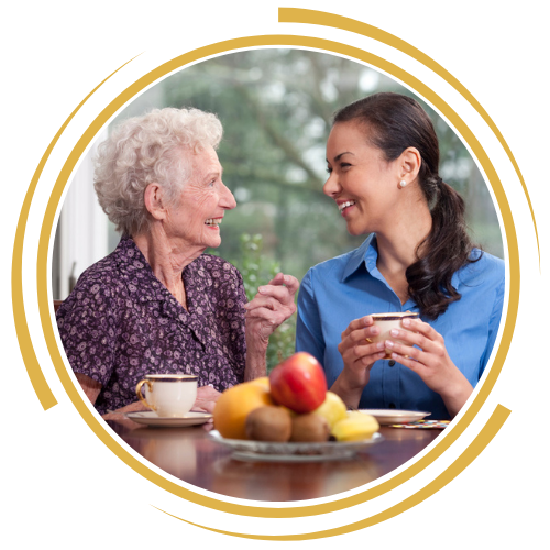 An elderly woman and a young woman smiling and talking at a table with a fruit bowl and cups of tea, enjoying a moment together.