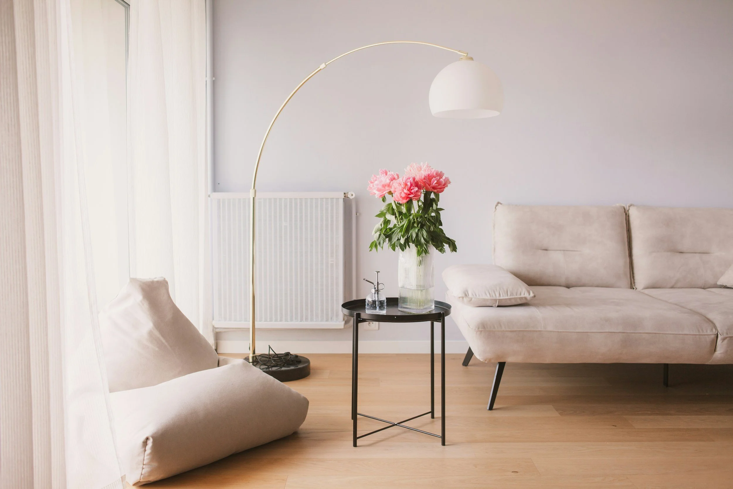 Living room with beige sofa and floor cushions, a black side table holding a large glass vase with pink peonies, a small spray bottle, and a lamp with a white shade and a golden arc, with light wood flooring and white walls.