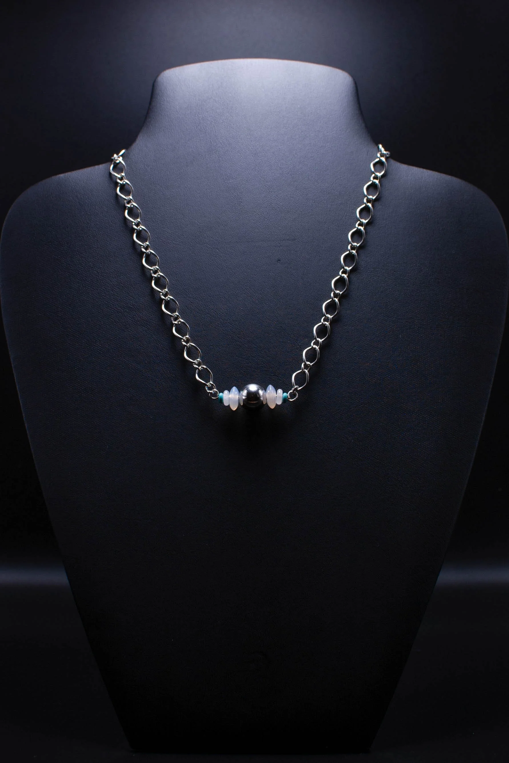 Silver chain necklace with a large central silver bead, flanked by smaller white, green, and blue beads, displayed on a black jewelry bust.