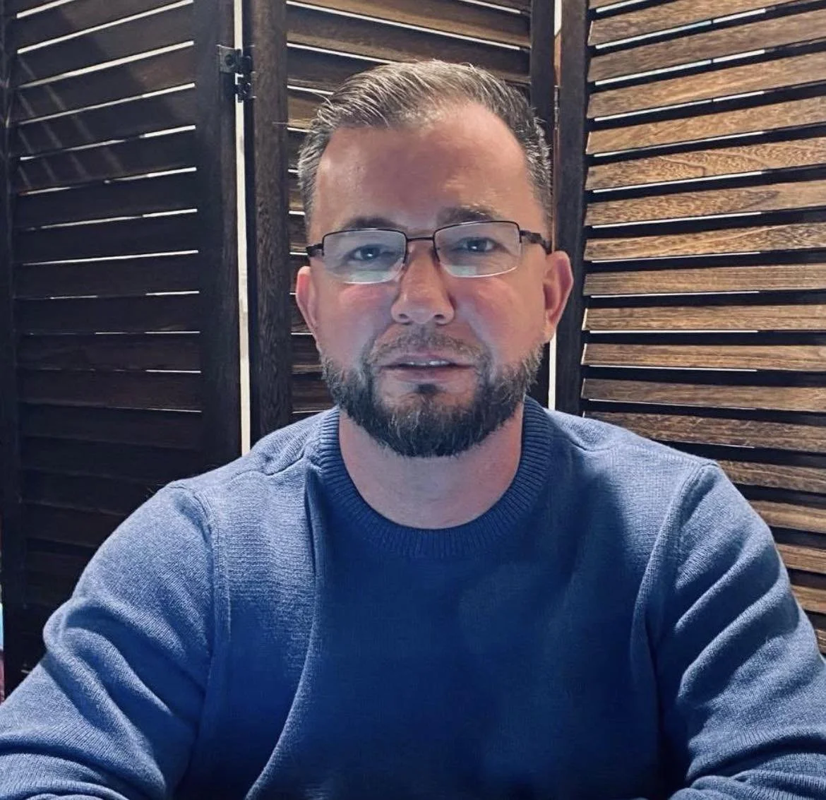 A man with glasses and a beard sitting in front of wooden slatted dividers in a indoor setting.