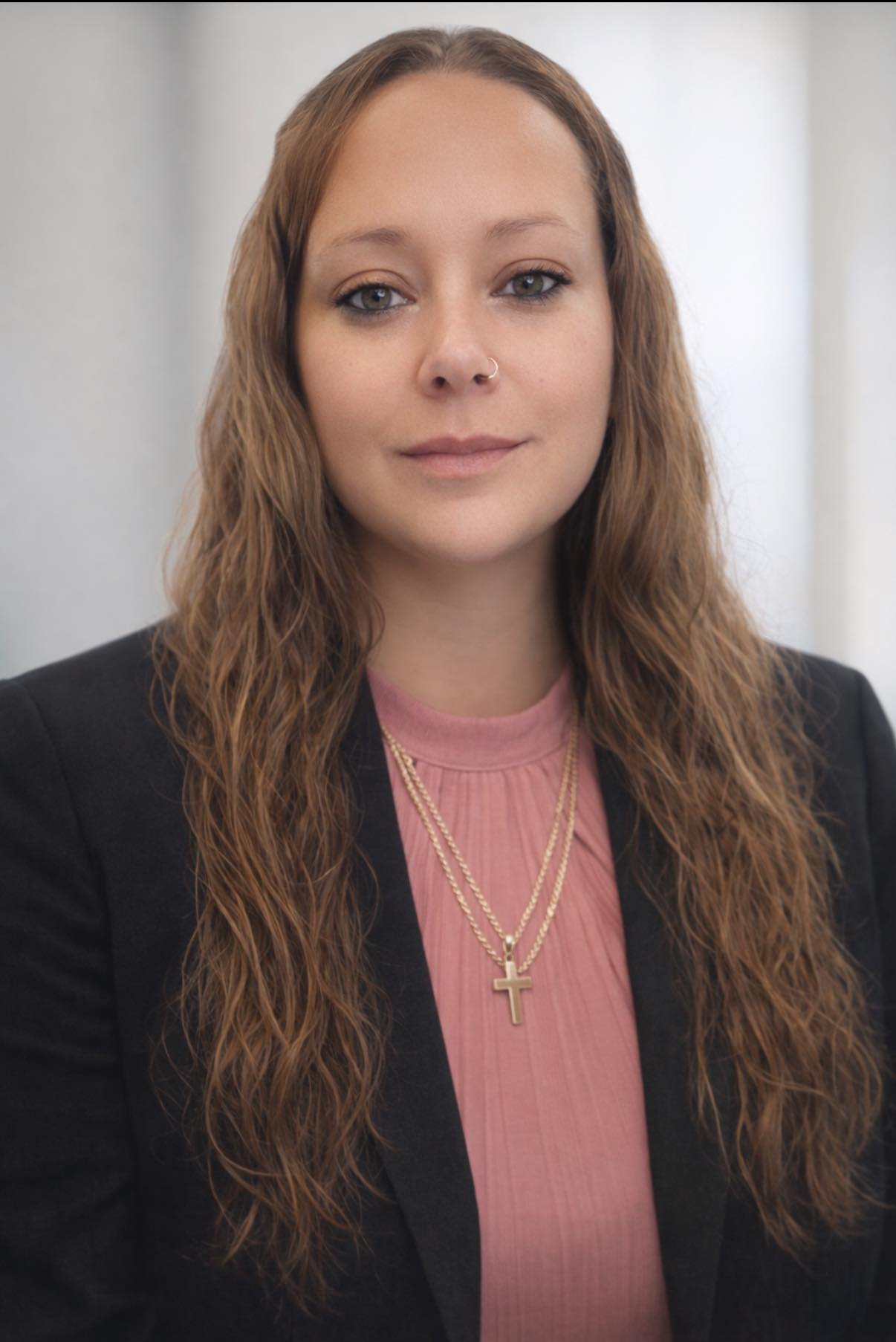 A woman with long wavy brown hair, wearing a black blazer over a pink top, gold necklaces with cross pendants, and small hoop nose ring, standing against a plain white background.