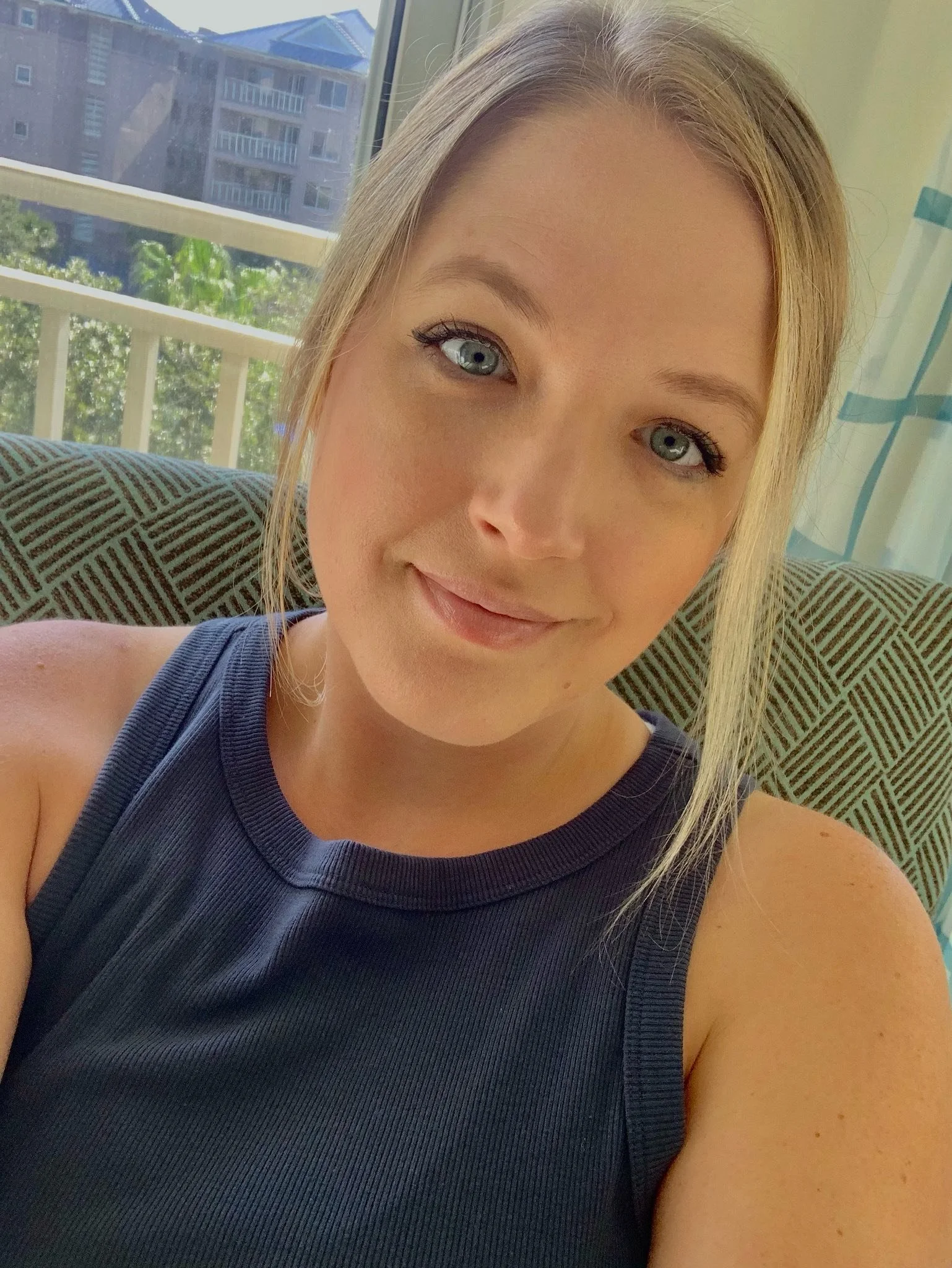 A woman with blonde hair, blue eyes, and wearing a sleeveless dark blue top, sitting indoors in front of a window with a view of a building and greenery outside.