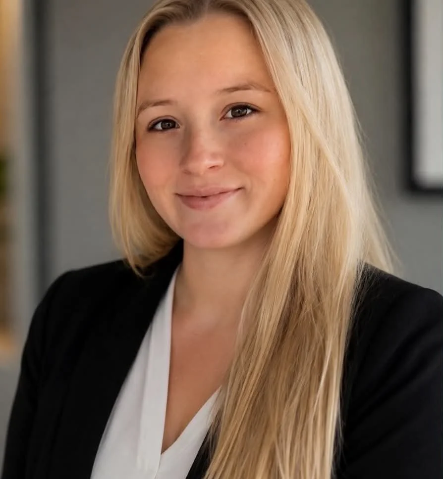 A young woman with long blonde hair smiling at the camera, wearing a black blazer and white blouse, with a blurred office background.