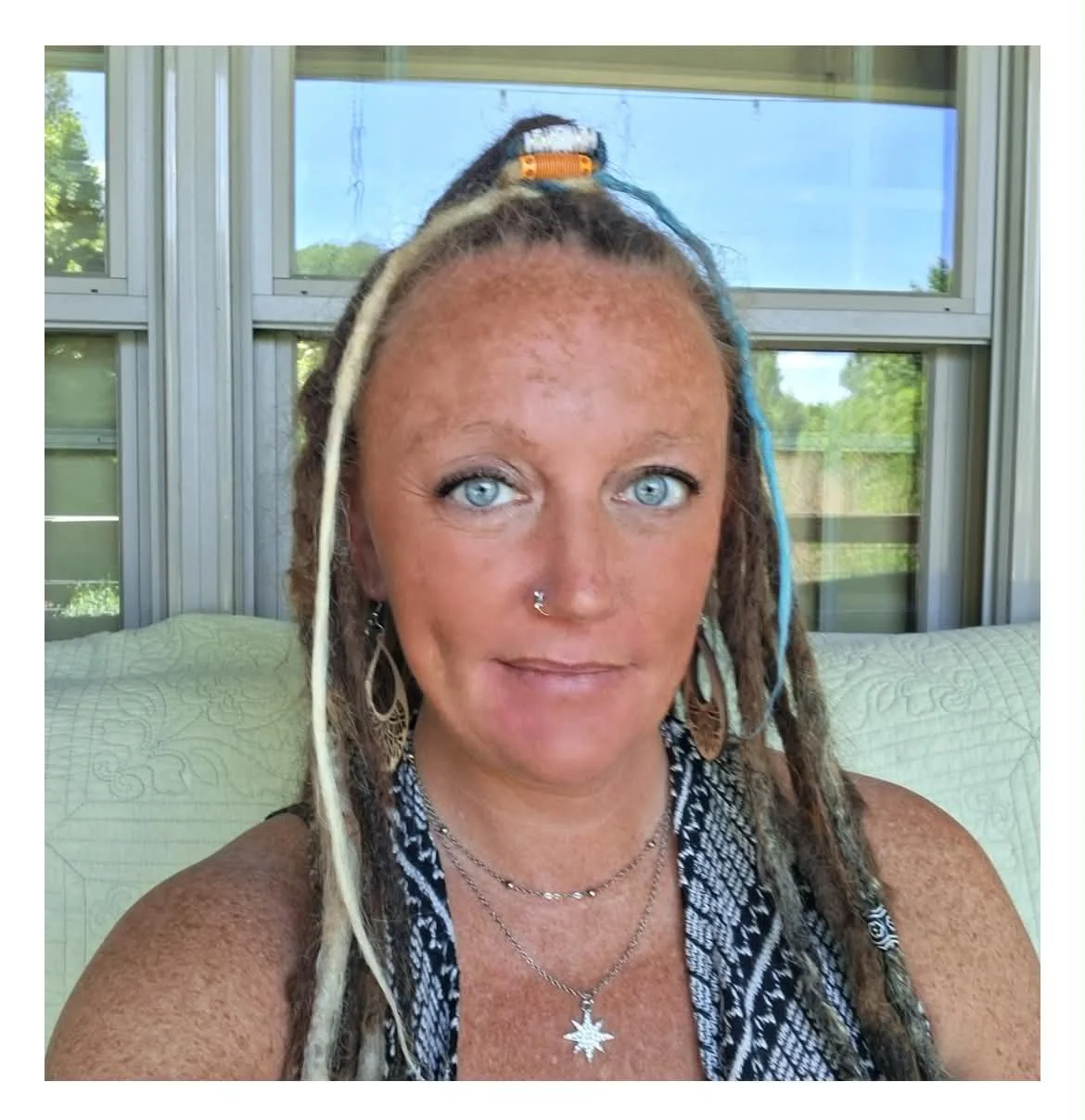 A woman with light skin and blue eyes, wearing earrings, necklaces, and with blonde dreadlocks featuring some colored strands or accessories, sitting on a screened porch with a quilted cushion behind her, and a background of trees and buildings outside.